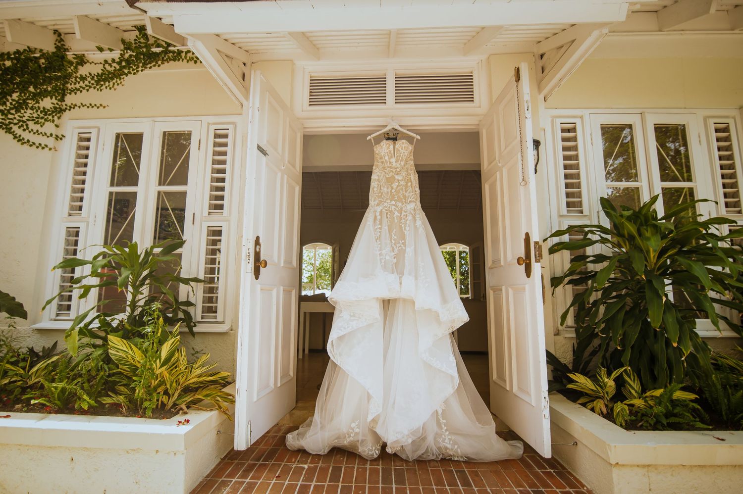 A wedding dress hanging in a bright, airy room with tropical plants and white decor.