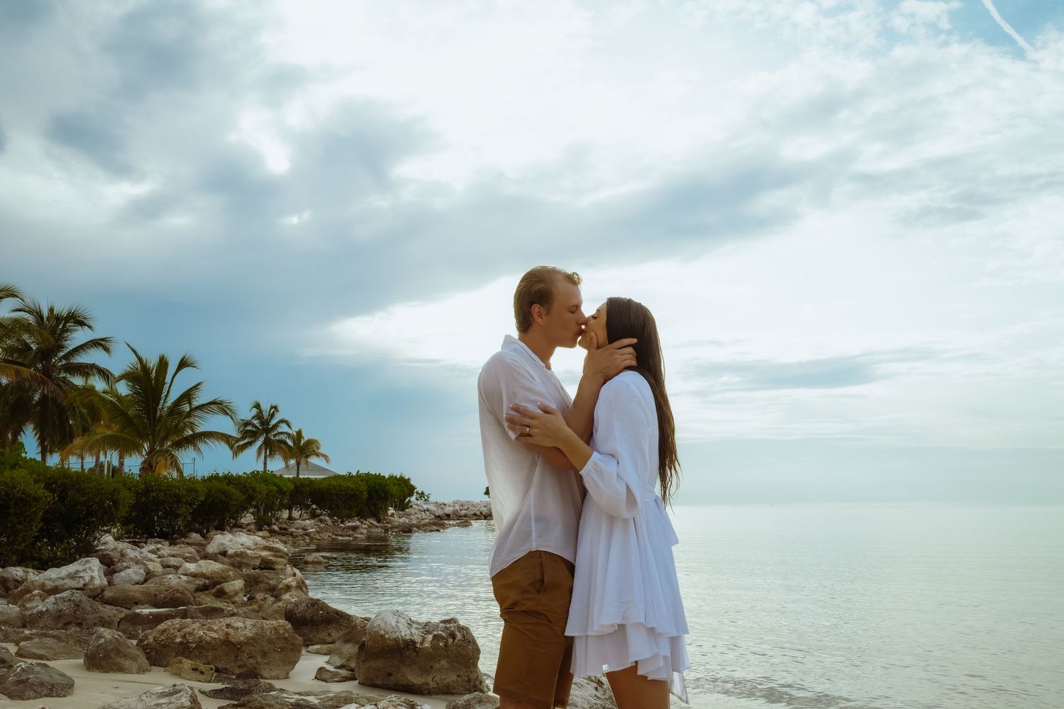 A couple shares a romantic moment on rocky coastal cliffs with palm trees and ocean backdrop.