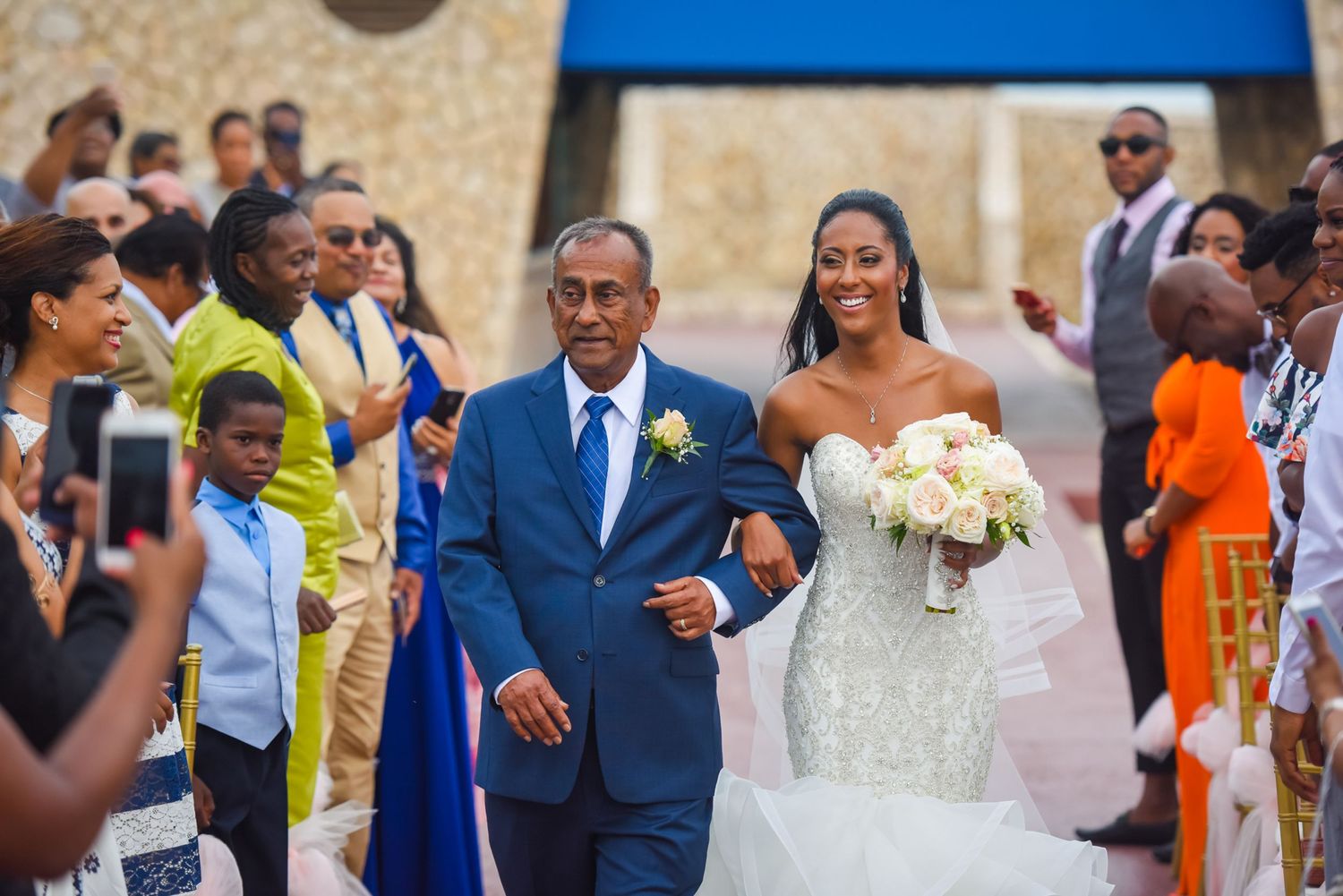 Wedding processional with guests capturing photos along stone pathway.