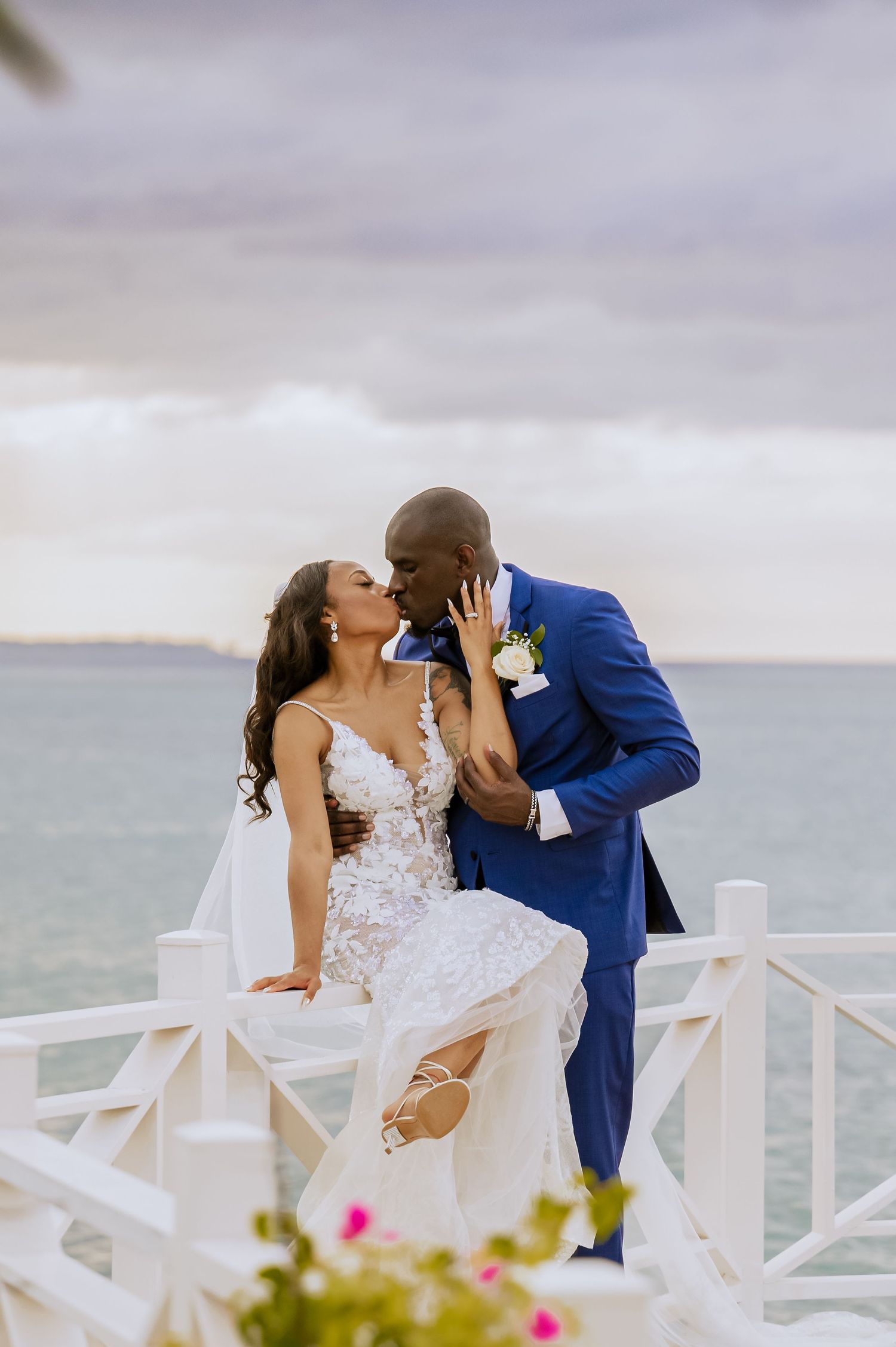 A romantic wedding couple shares a tender kiss on a waterfront deck with white railings at sunset.