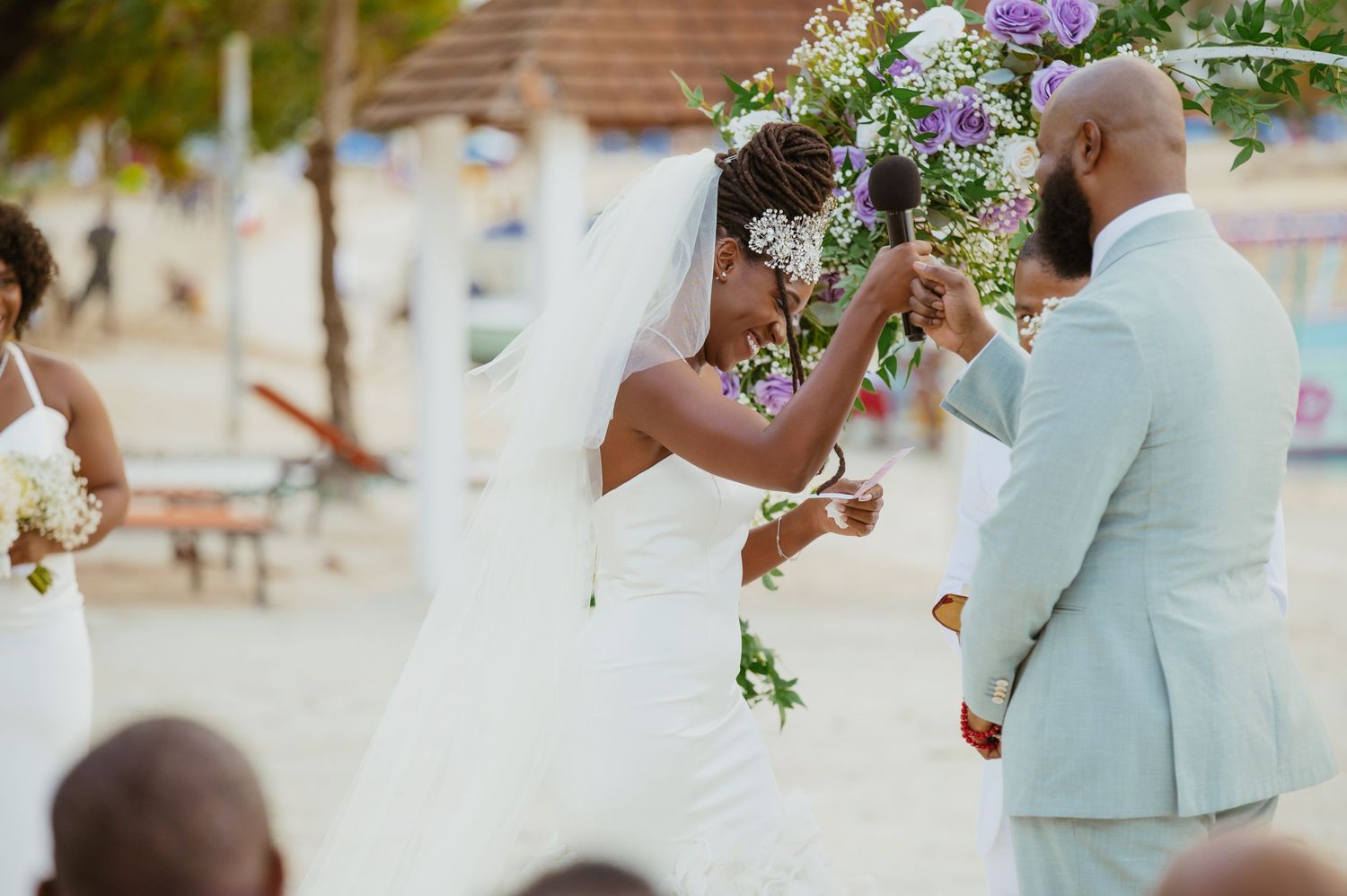 Beachside wedding ceremony with purple floral decorations and guests in light-colored attire.