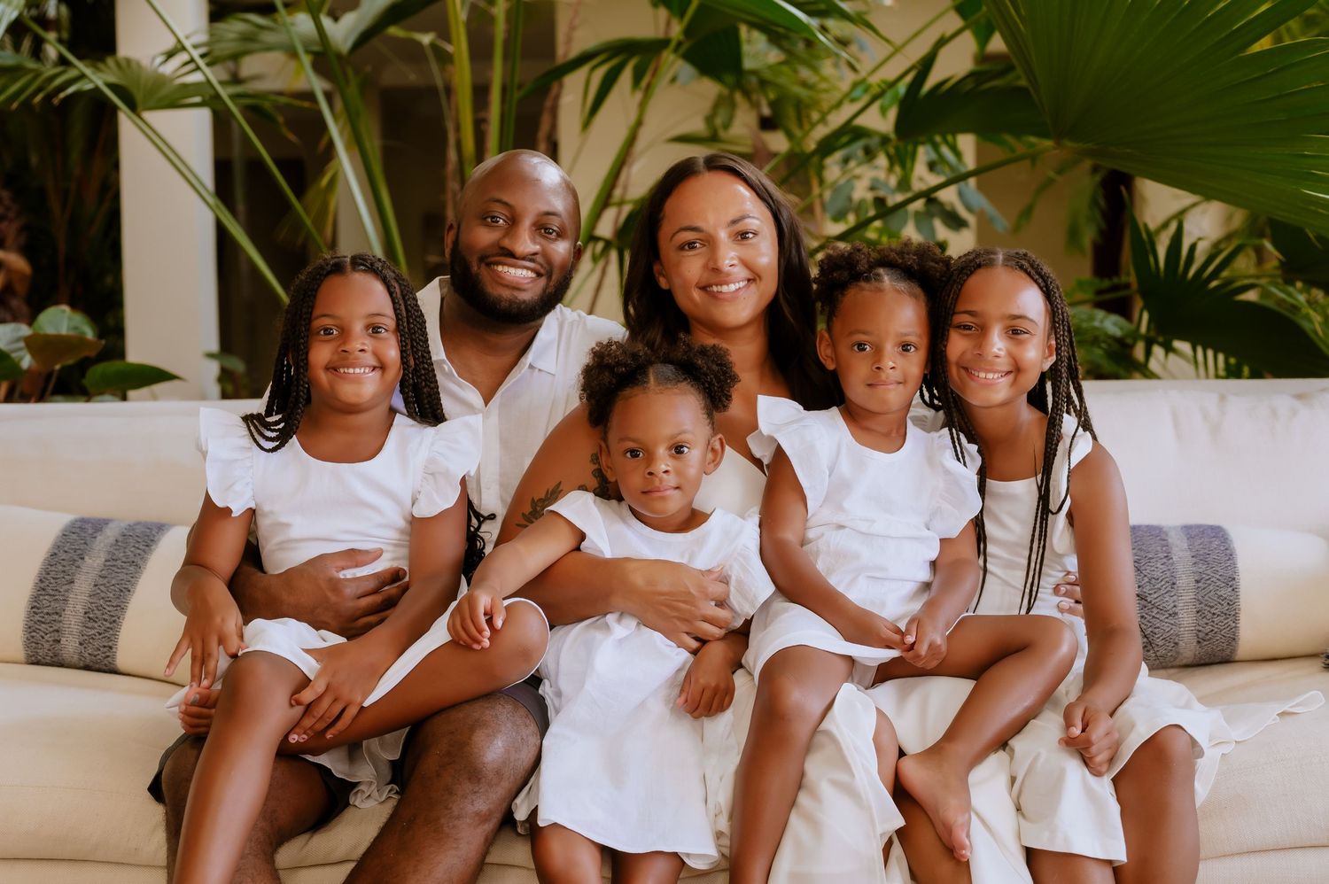 Family of six wearing matching white outfits smile while sitting together on a gray couch.