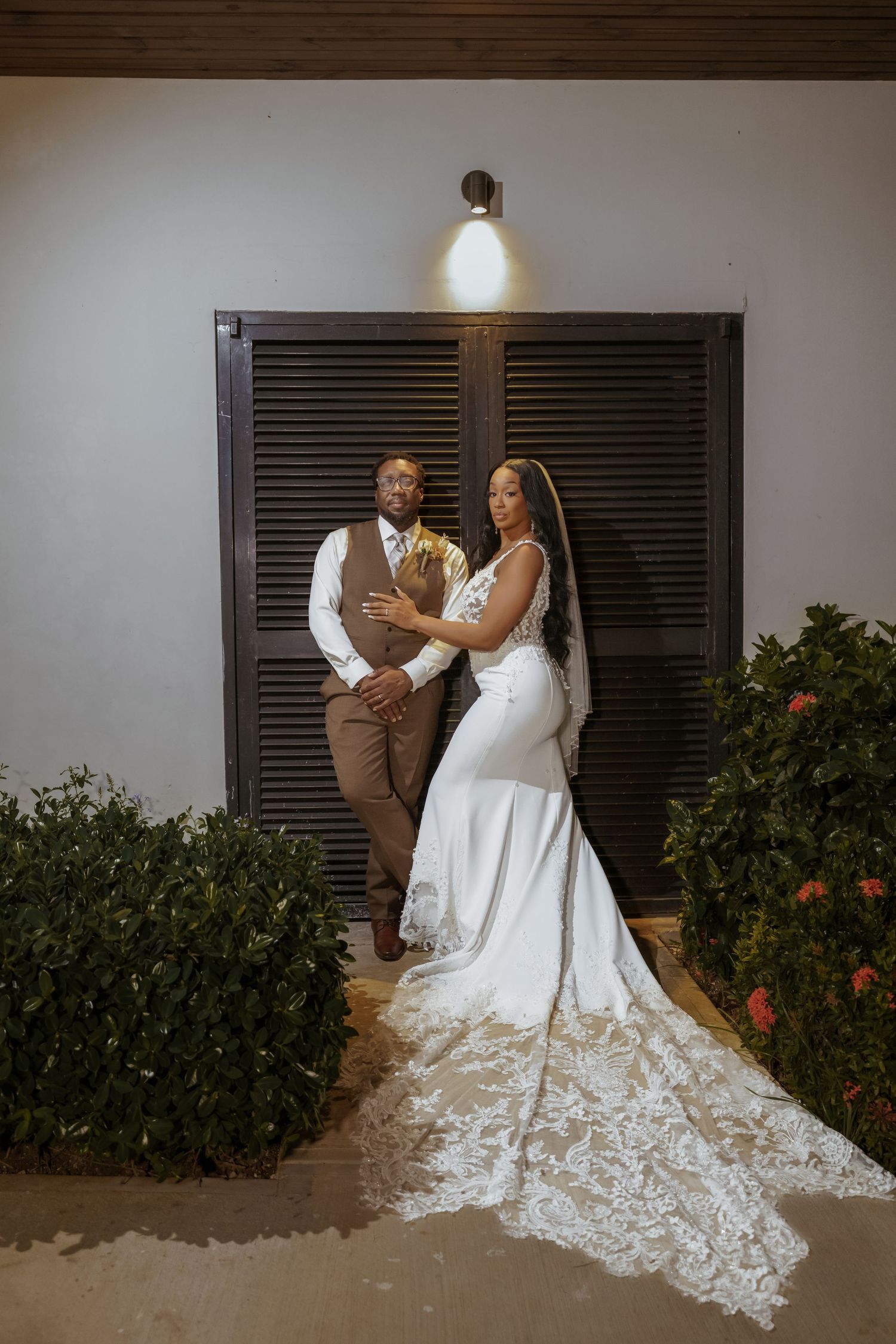 Wedding couple poses against dark shutters with bride's lace train flowing beautifully.