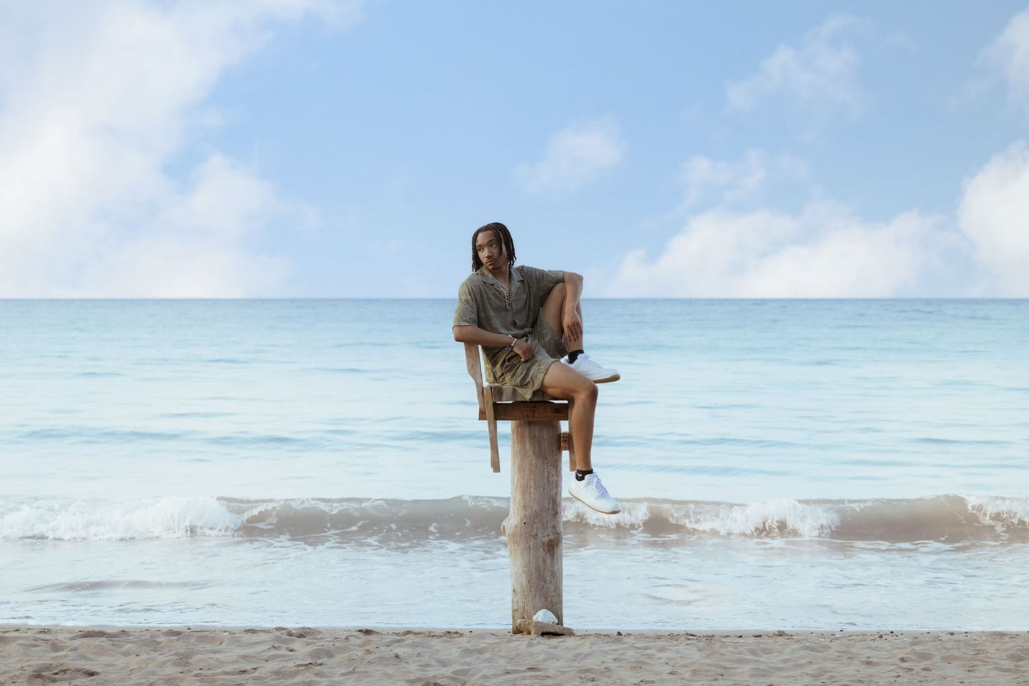 A figure sits on a wooden post at the beach with turquoise waters and cloudy skies in the background.