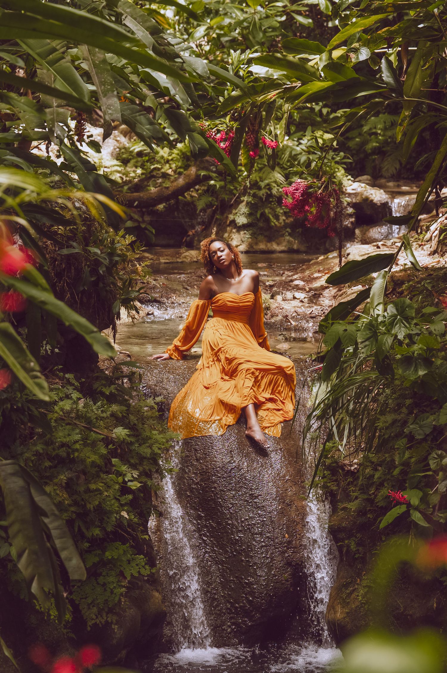 Woman in orange dress poses playfully near a tropical waterfall surrounded by lush greenery.
