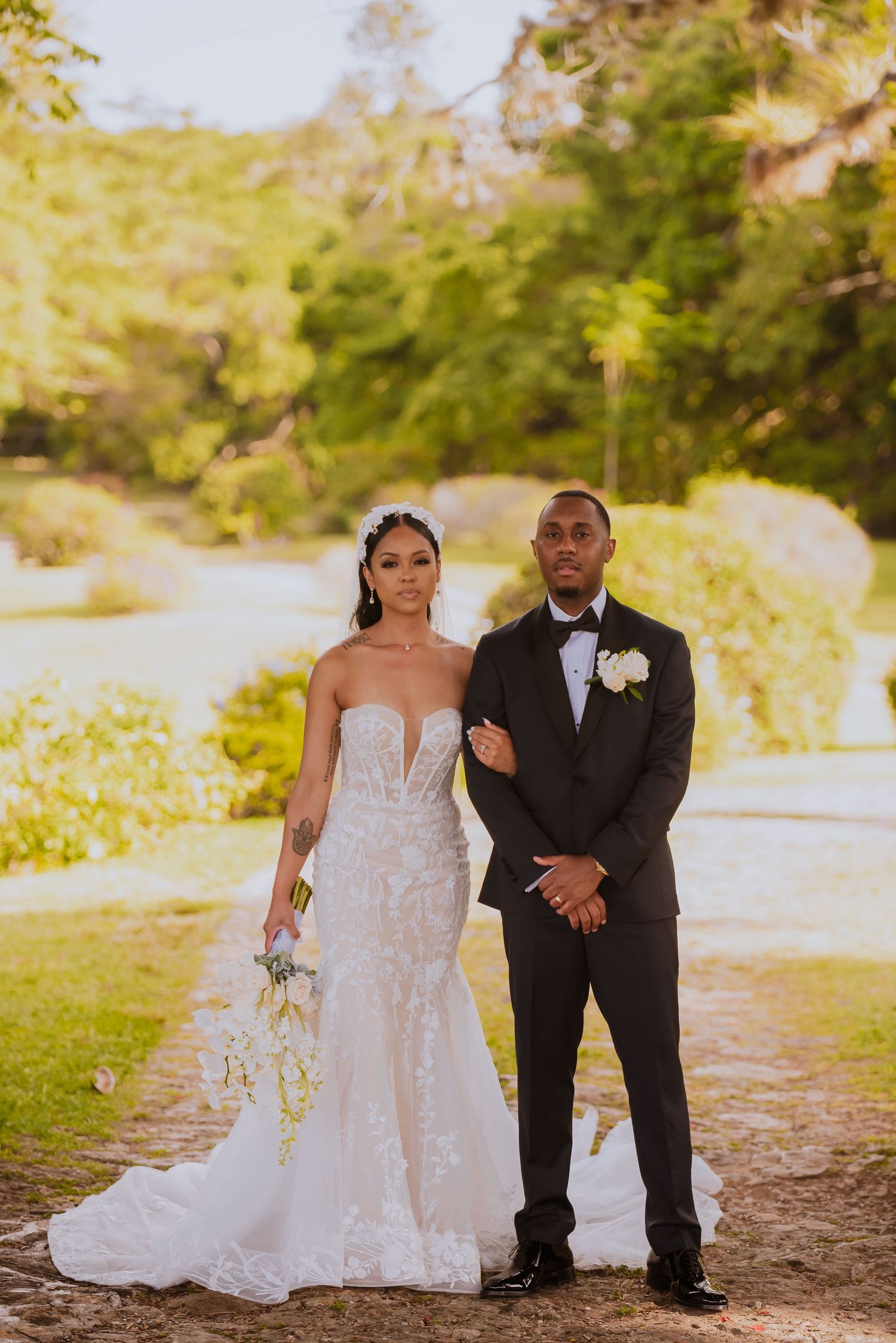 A bride in a flowing white gown and groom in black suit pose together in a garden setting.
