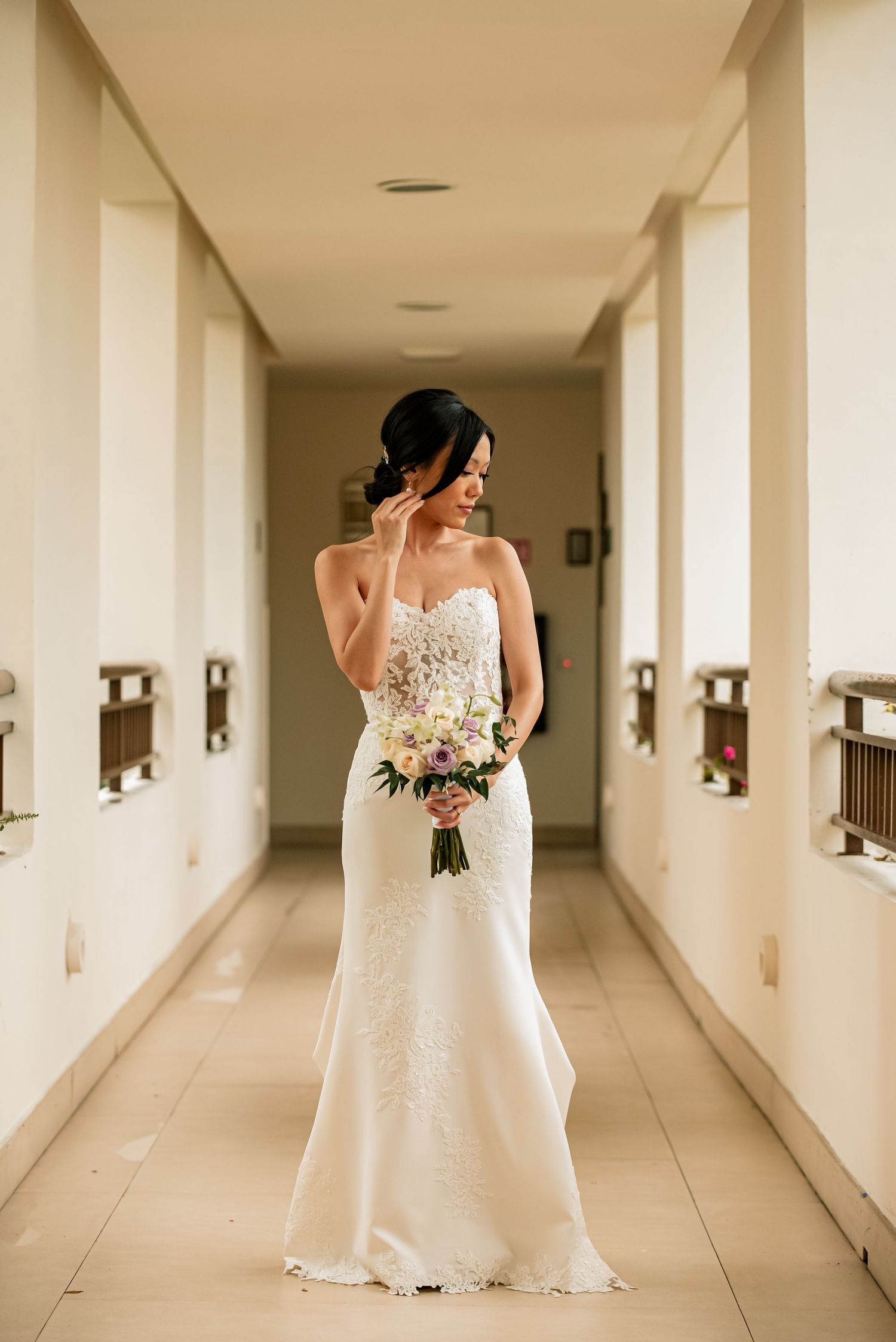 A bride in a white lace wedding dress holds a bouquet while standing in a long corridor.