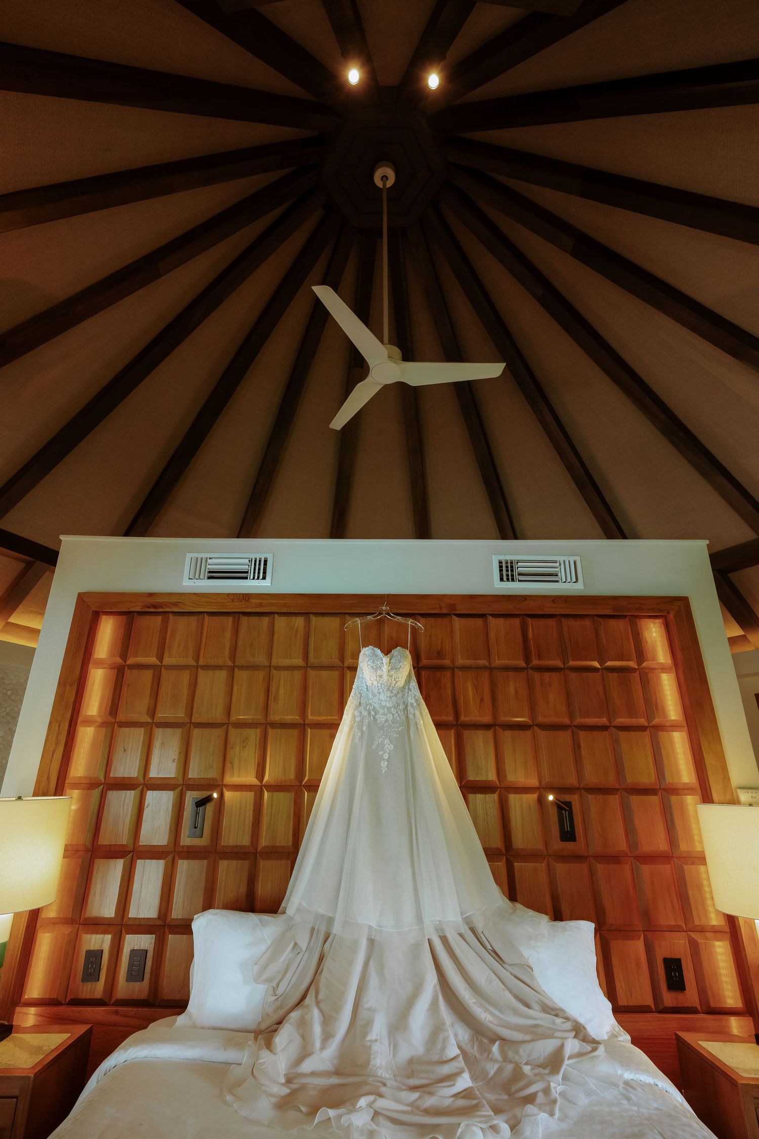 An elegant white wedding dress hanging in a room with dramatic wooden beam ceiling and lighting.