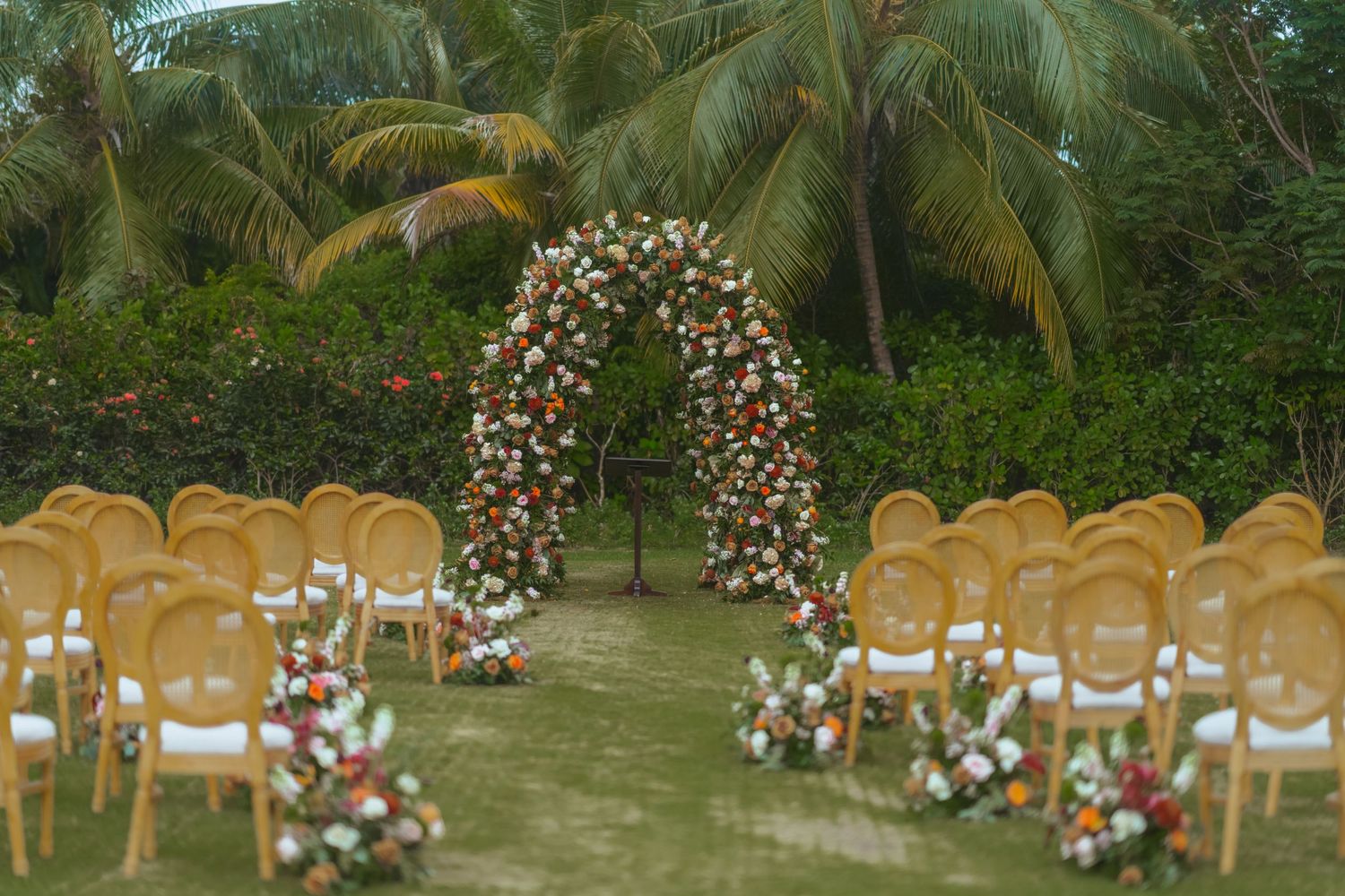 Outdoor wedding ceremony setup with wooden chairs and floral arches under tropical palm trees. Luxury wedding photography at Half Moon