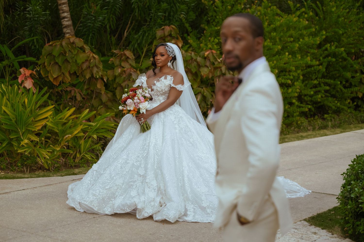 Bride in white gown and groom in cream suit pose together in lush garden setting.