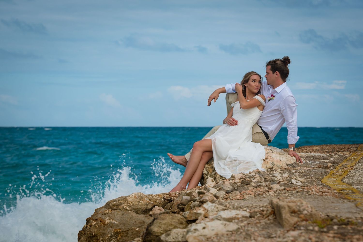 Wedding couple sitting on rocky coastline as waves crash dramatically in background.