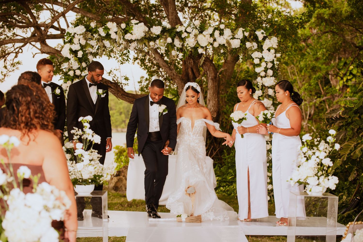 Wedding party gathered outdoors under trees with white floral decorations for a ceremony.