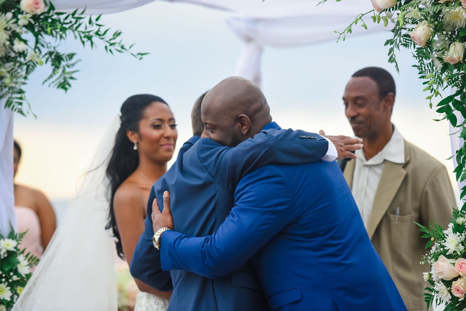 Emotional embrace shared during outdoor wedding ceremony under floral decorations and white tent.