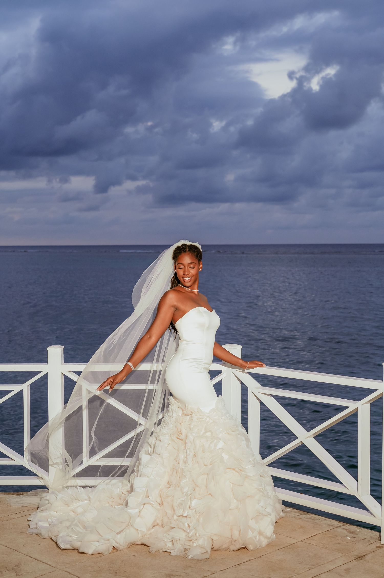 A bride in a flowing white dress poses dramatically on a white railing overlooking the ocean under stormy skies.