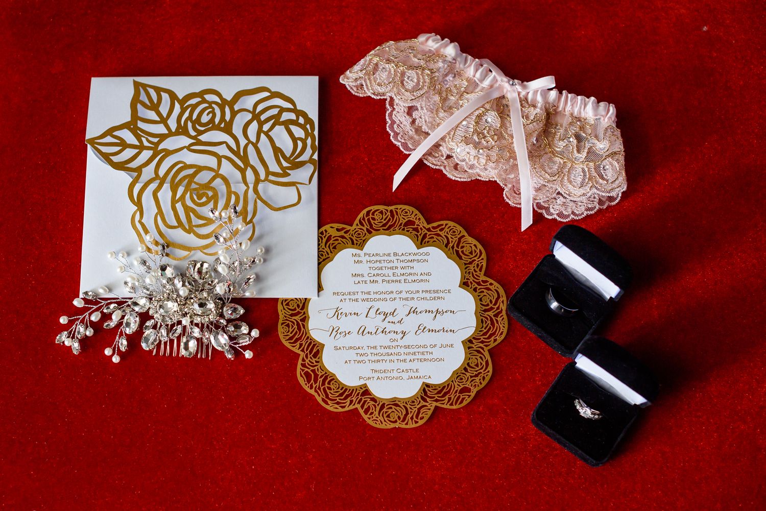 Wedding accessories including a lace garter, gold floral invitation, and black shoes arranged on red fabric.