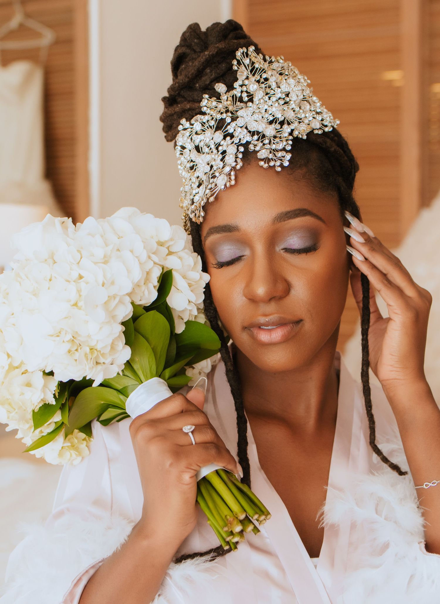 Bride wearing ornate crystal headpiece holds white bouquet with closed eyes in serene moment.