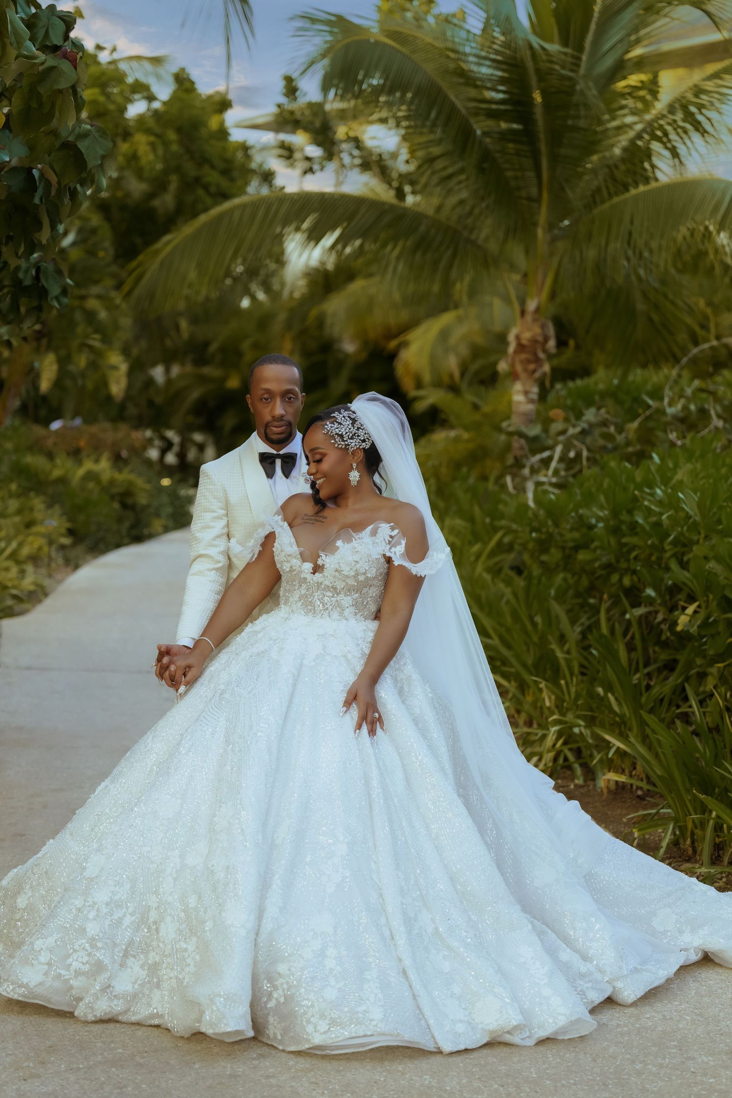 Couple in elegant white wedding attire posing together on a tropical beach with palm trees.