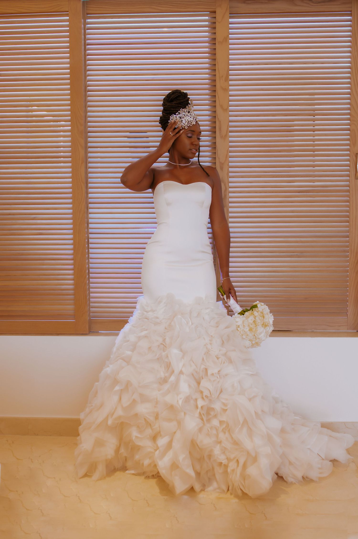 Bride in strapless ruffled wedding gown poses against wooden shutters with bouquet.