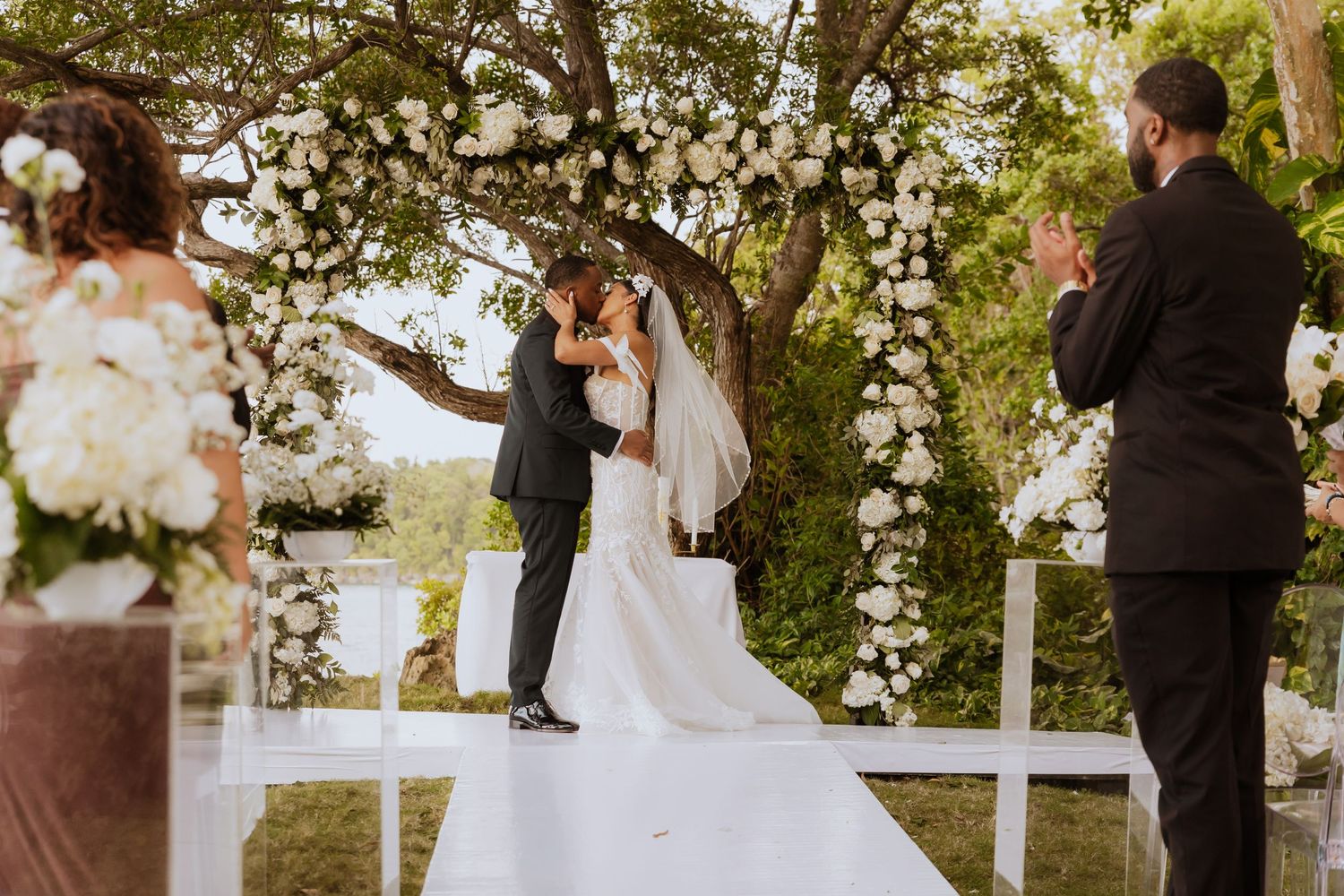 Wedding ceremony taking place under trees with white flower arrangements and wooden arch.