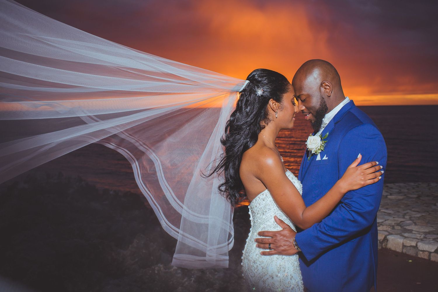 Newlyweds kiss under flowing white veil with dramatic sunset colors over ocean backdrop.