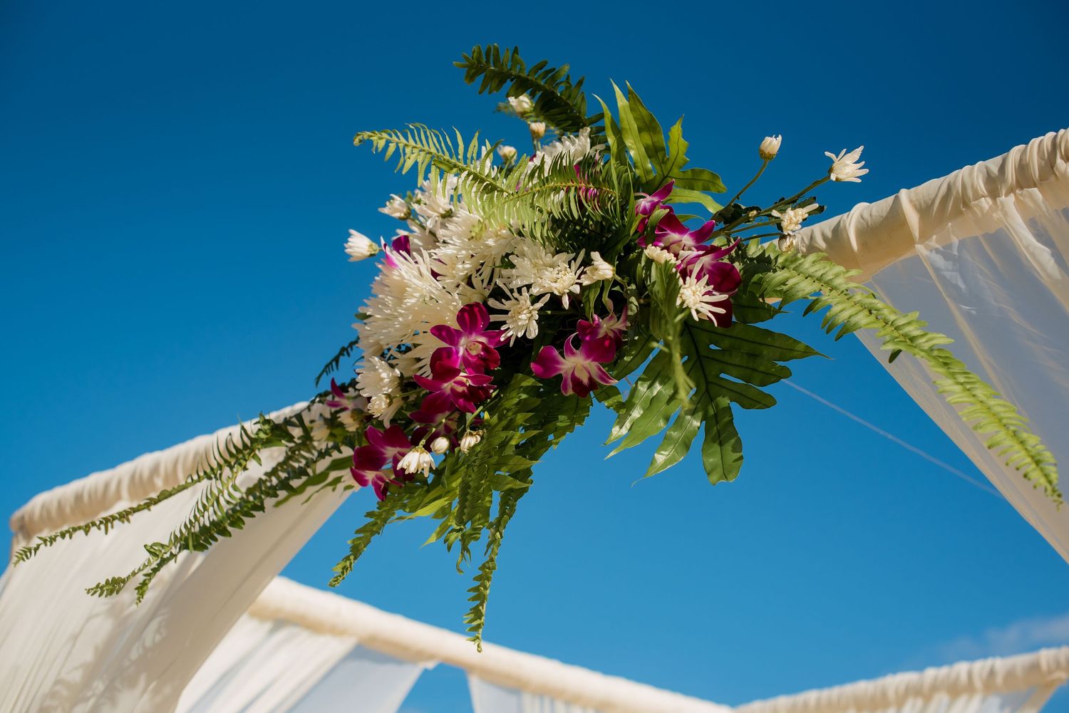 Beach wedding arch decorated with tropical flowers against bright blue sky.
