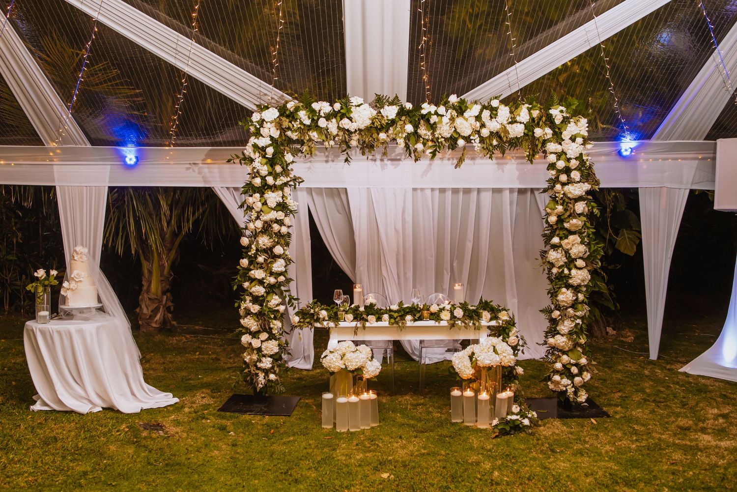 Elegant outdoor wedding setup with floral arch, white drapes and twinkling lights creating a romantic evening ambiance.