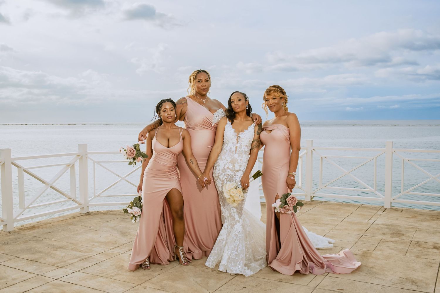 Bridal party in blush pink dresses poses with bride against ocean backdrop.