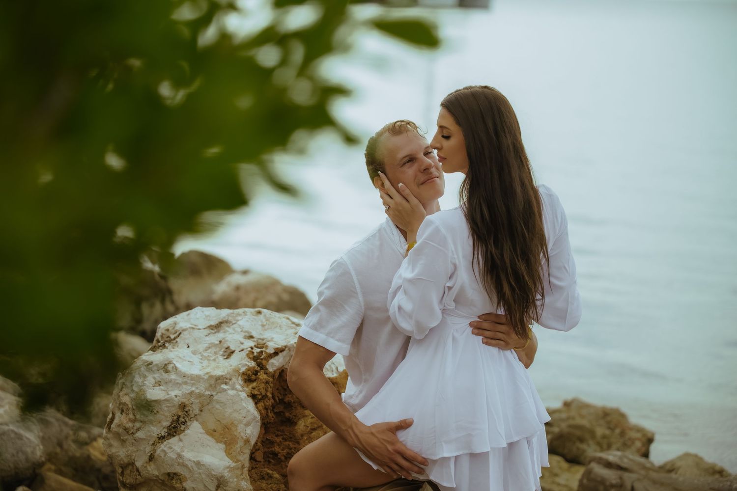 A couple in white clothing embraces while sitting on coastal rocks near calm waters.