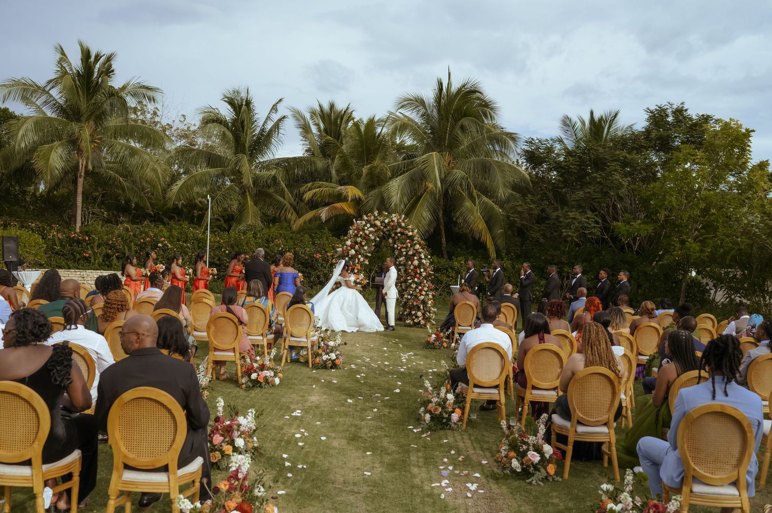 Outdoor wedding ceremony setup with rows of wooden chairs on a grassy lawn surrounded by palm trees.