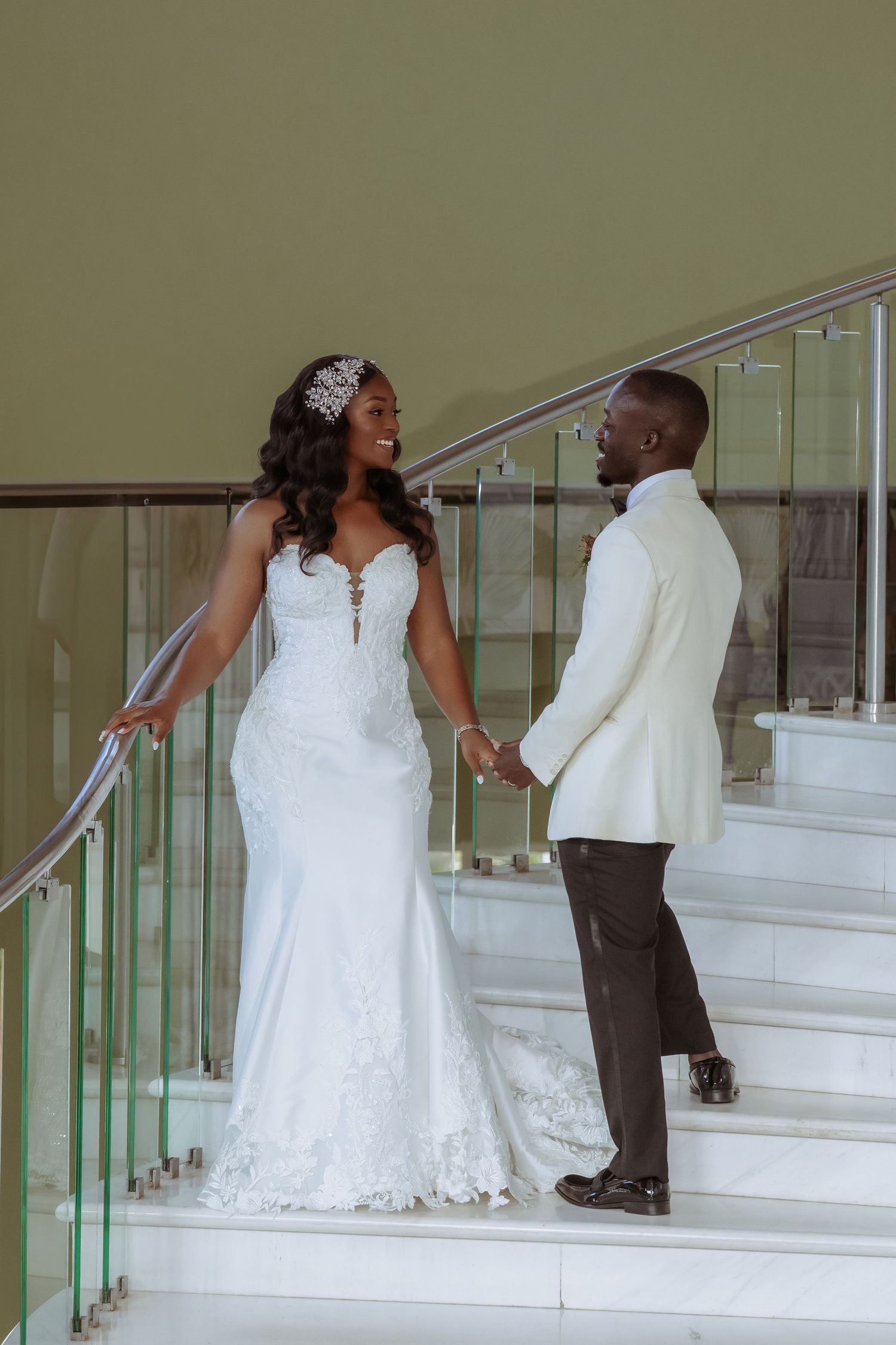 A wedding couple poses on elegant white stairs with glass railings.