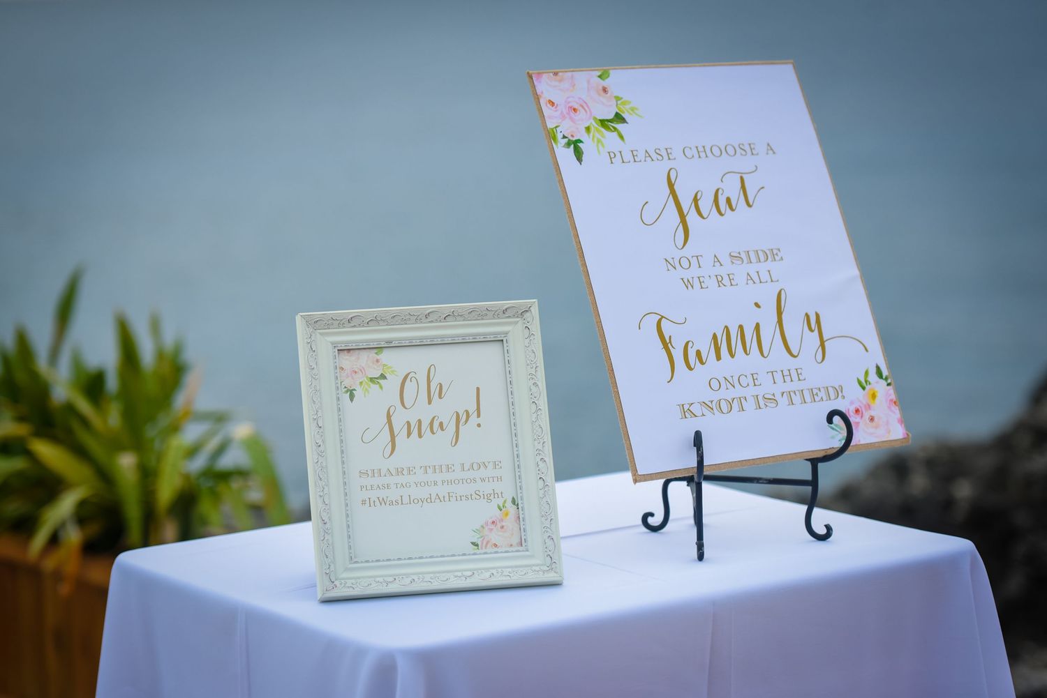 Wedding signage displayed on white table with floral accents and gold lettering.
