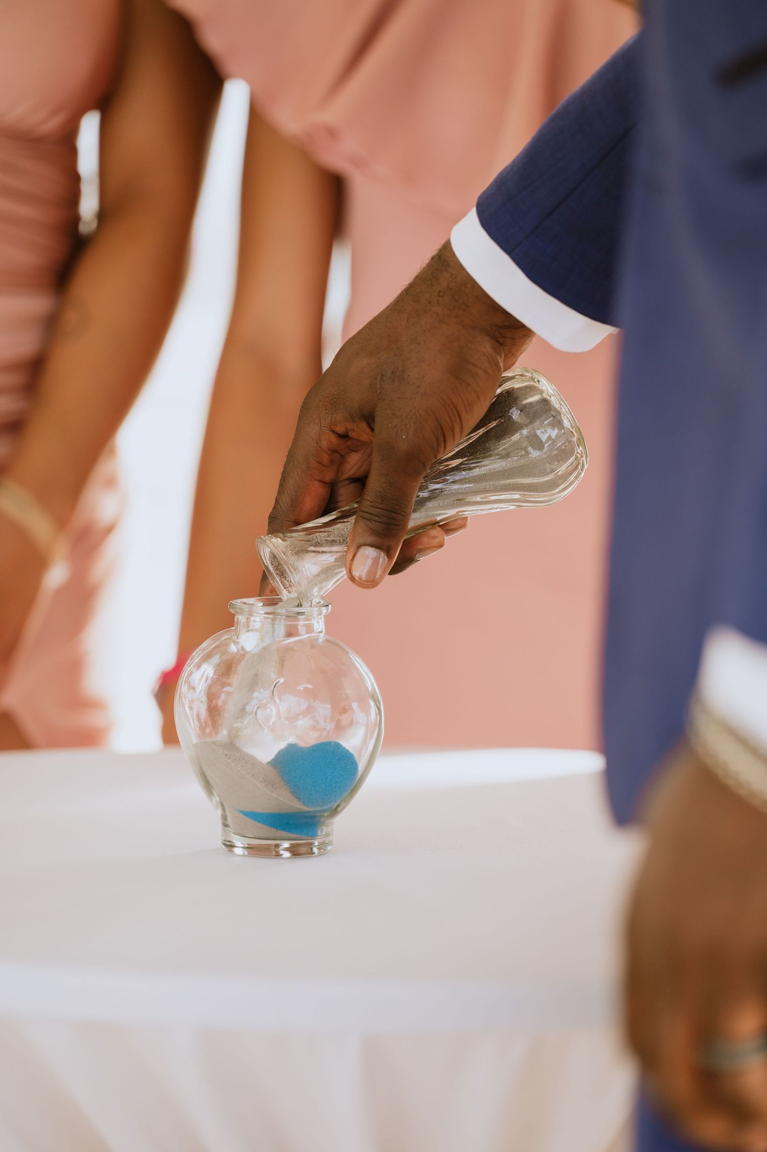 A hand pours blue sand into a decorative glass vessel during a wedding ceremony.