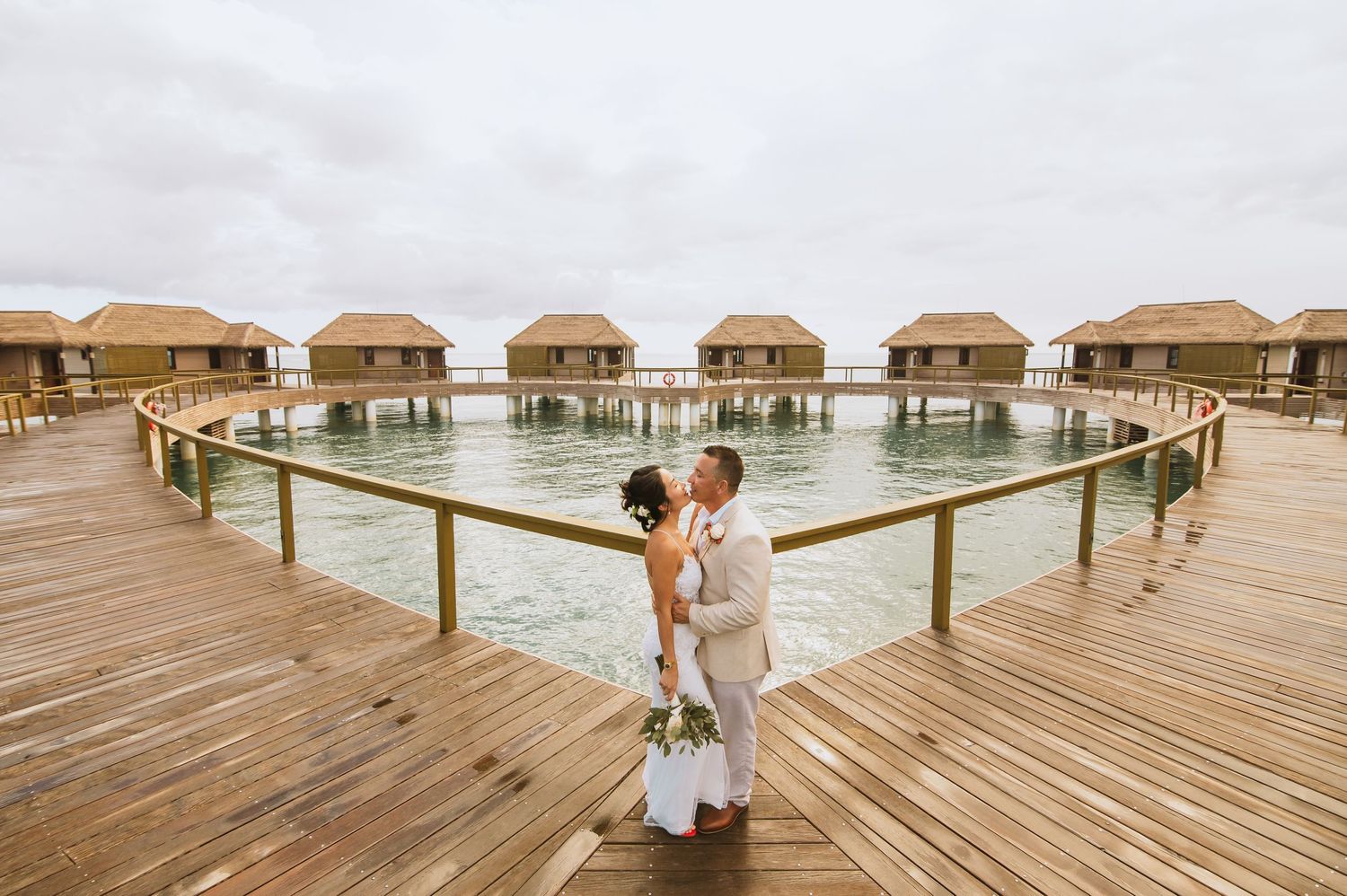 Couple standing on wooden boardwalk with overwater bungalows in the background at tropical resort.