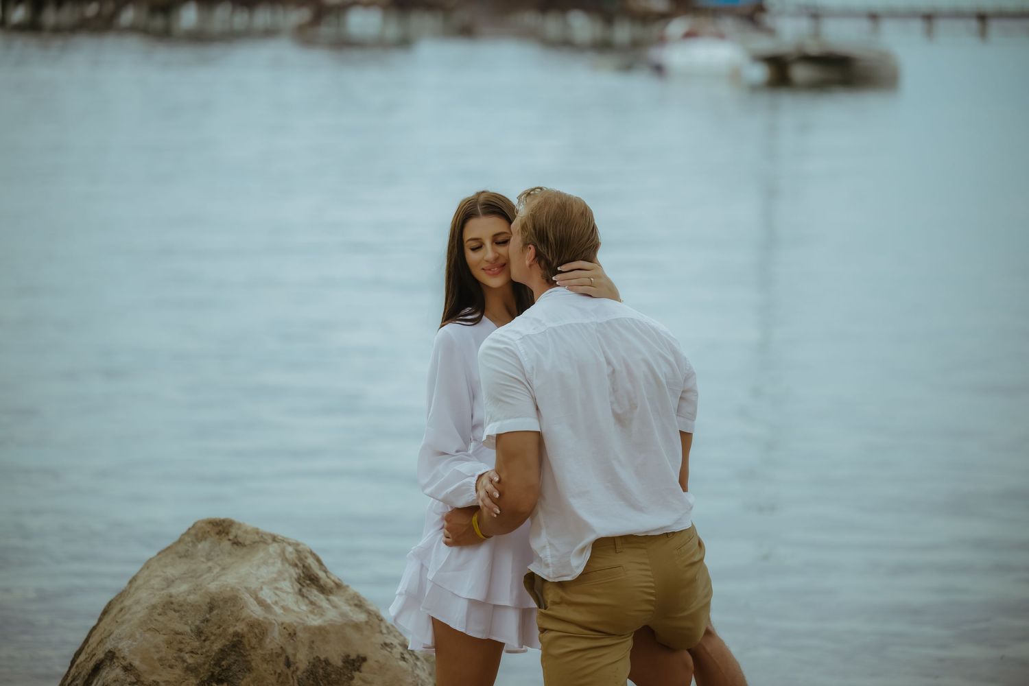 A couple shares a tender moment on rocky shoreline overlooking tranquil waters.