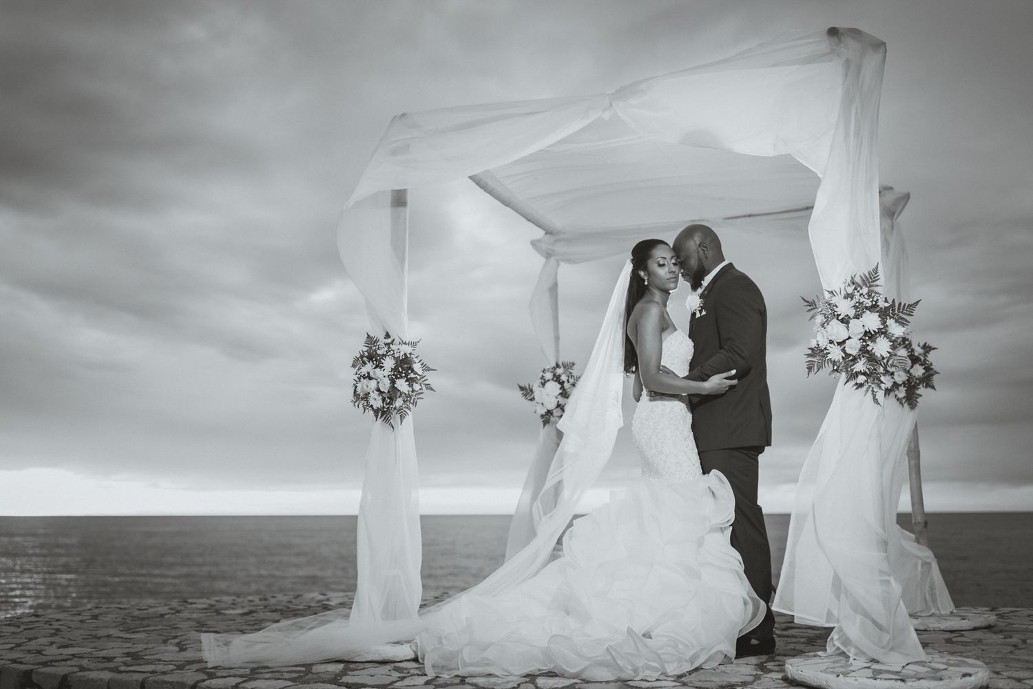 Black and white wedding photo capturing couple's romantic moment under seaside canopy.