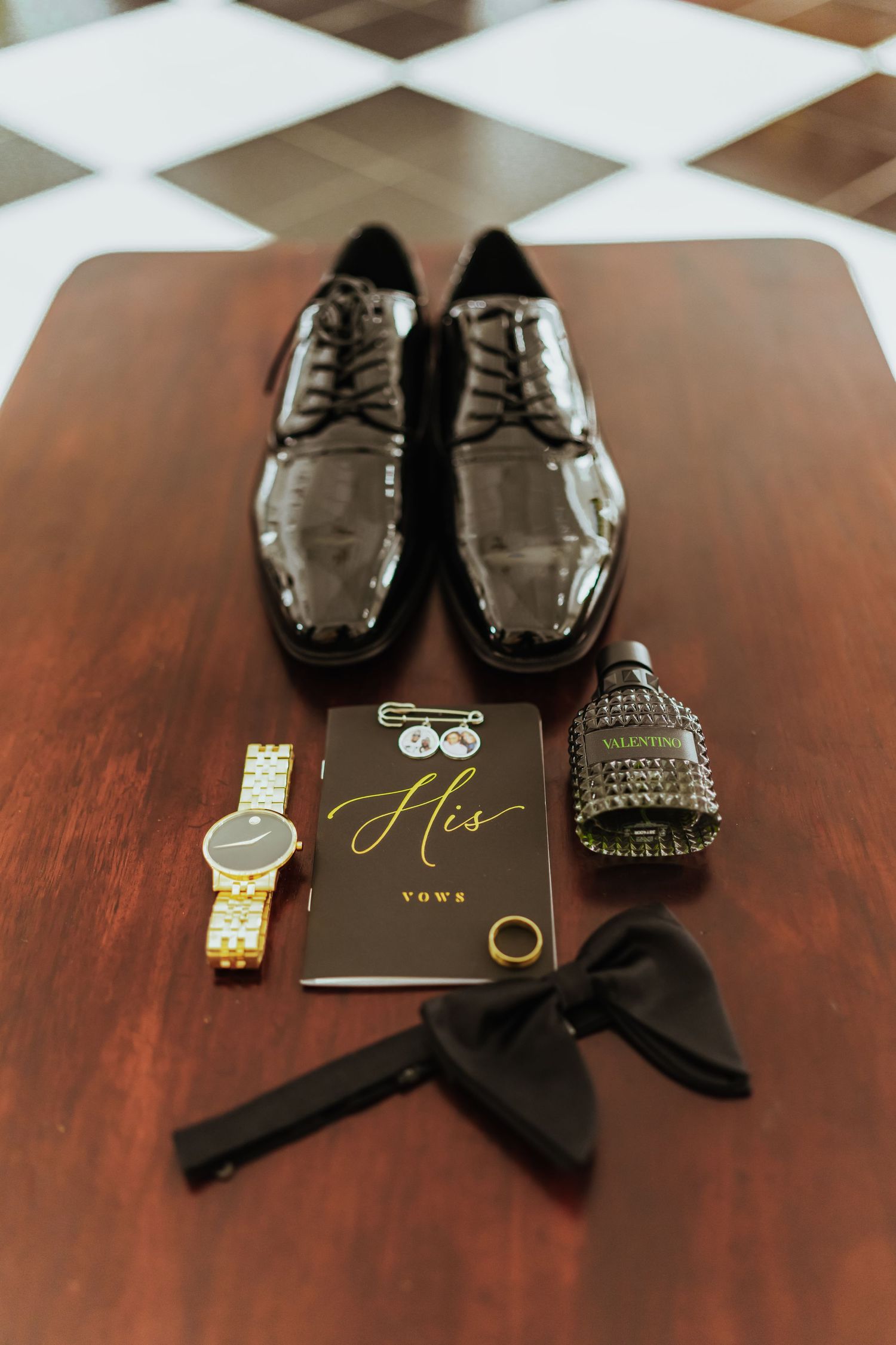 Wedding accessories including patent leather shoes, bow tie, watch and rings arranged on dark wooden surface.
