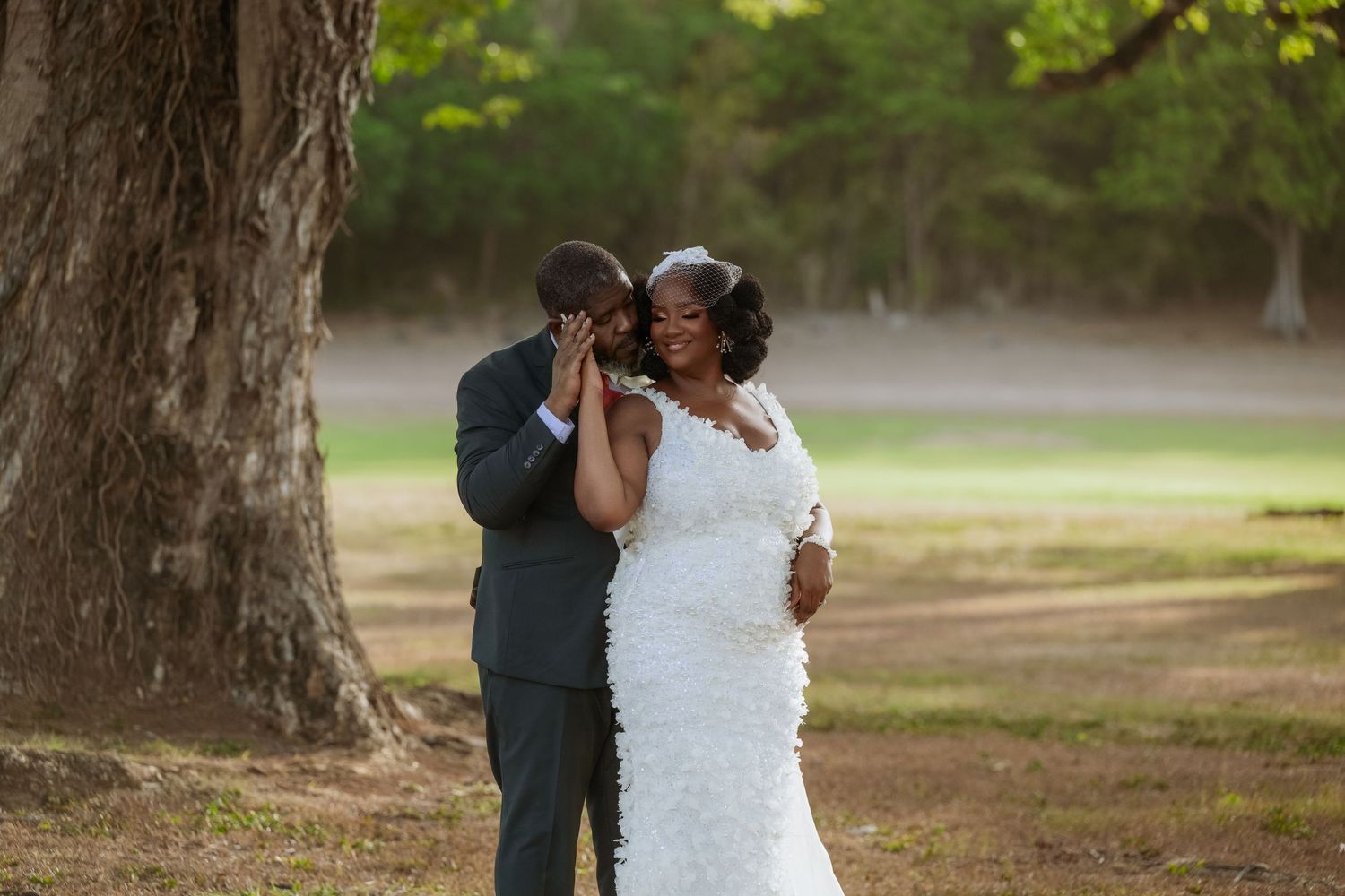A couple in wedding attire embrace under tall trees in a romantic outdoor setting.
