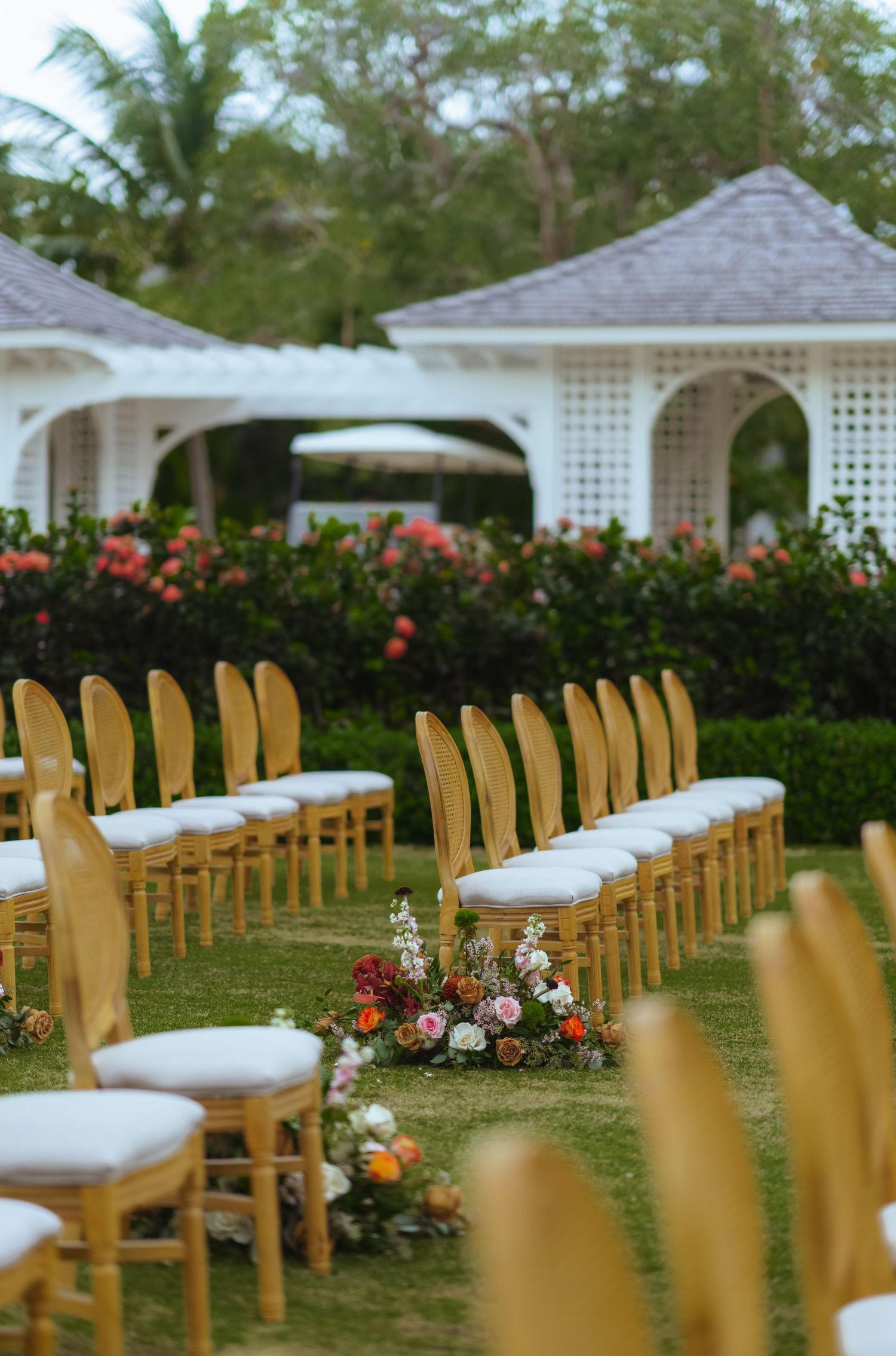 Outdoor wedding ceremony setup with wooden chairs and white cushions facing a white gazebo surrounded by tropical gardens.