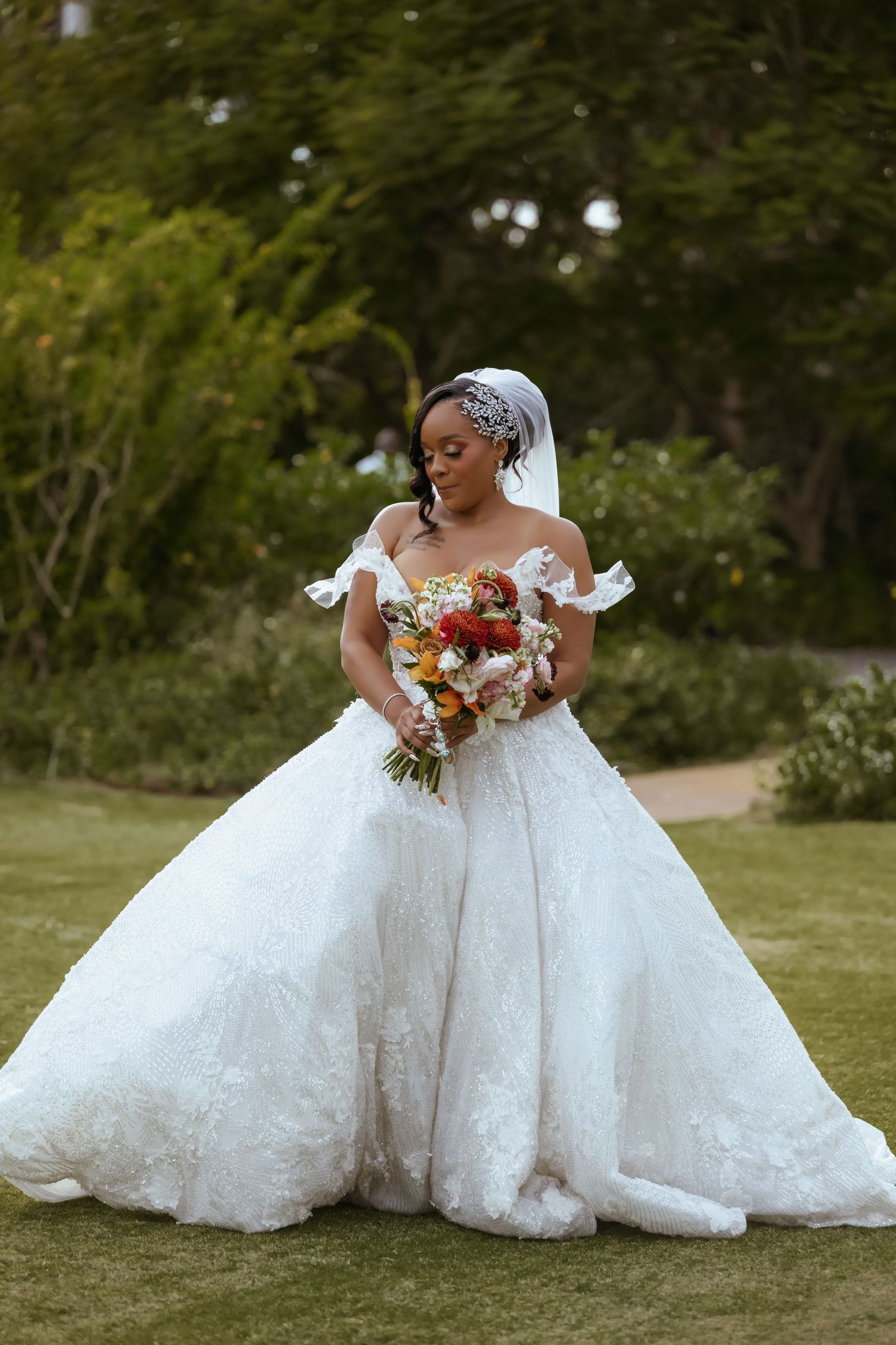 A bride in a luxurious ball gown wedding dress poses with a colorful bouquet in an outdoor garden setting.