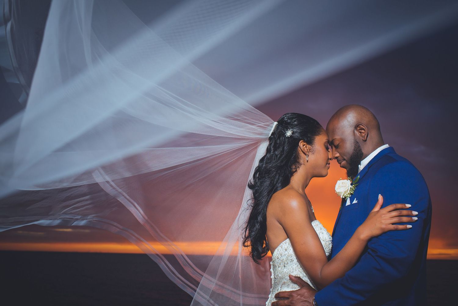 Wedding couple sharing intimate moment as bride's veil flows in ocean breeze at sunset.