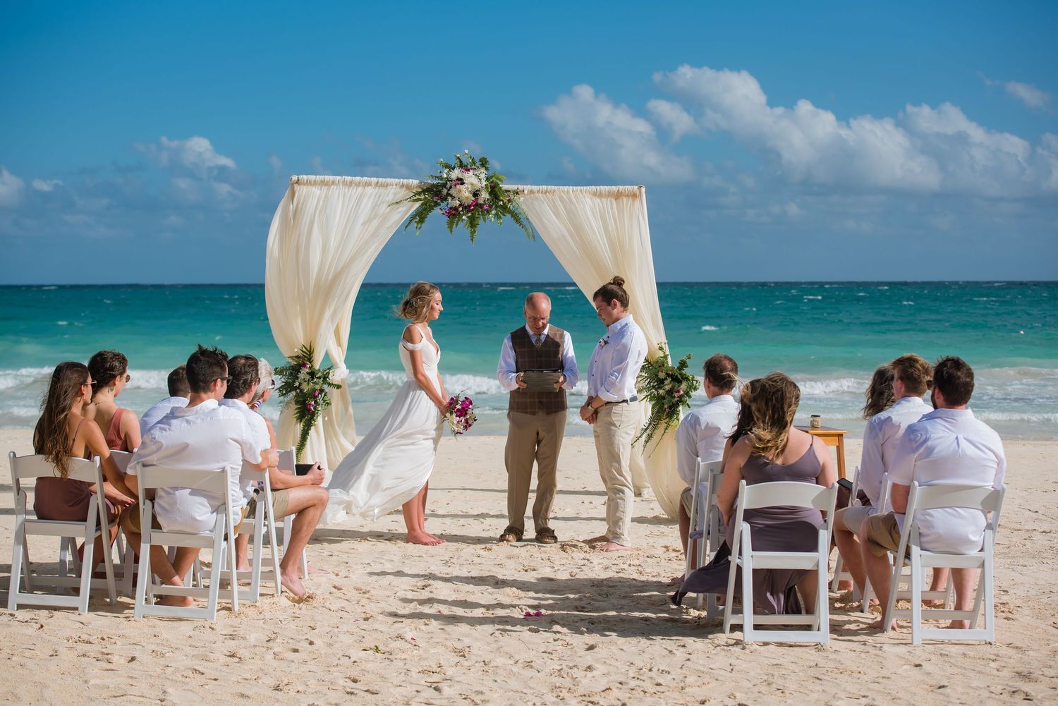 Wide view of beach wedding setup with white chairs and fabric arch along the shoreline.