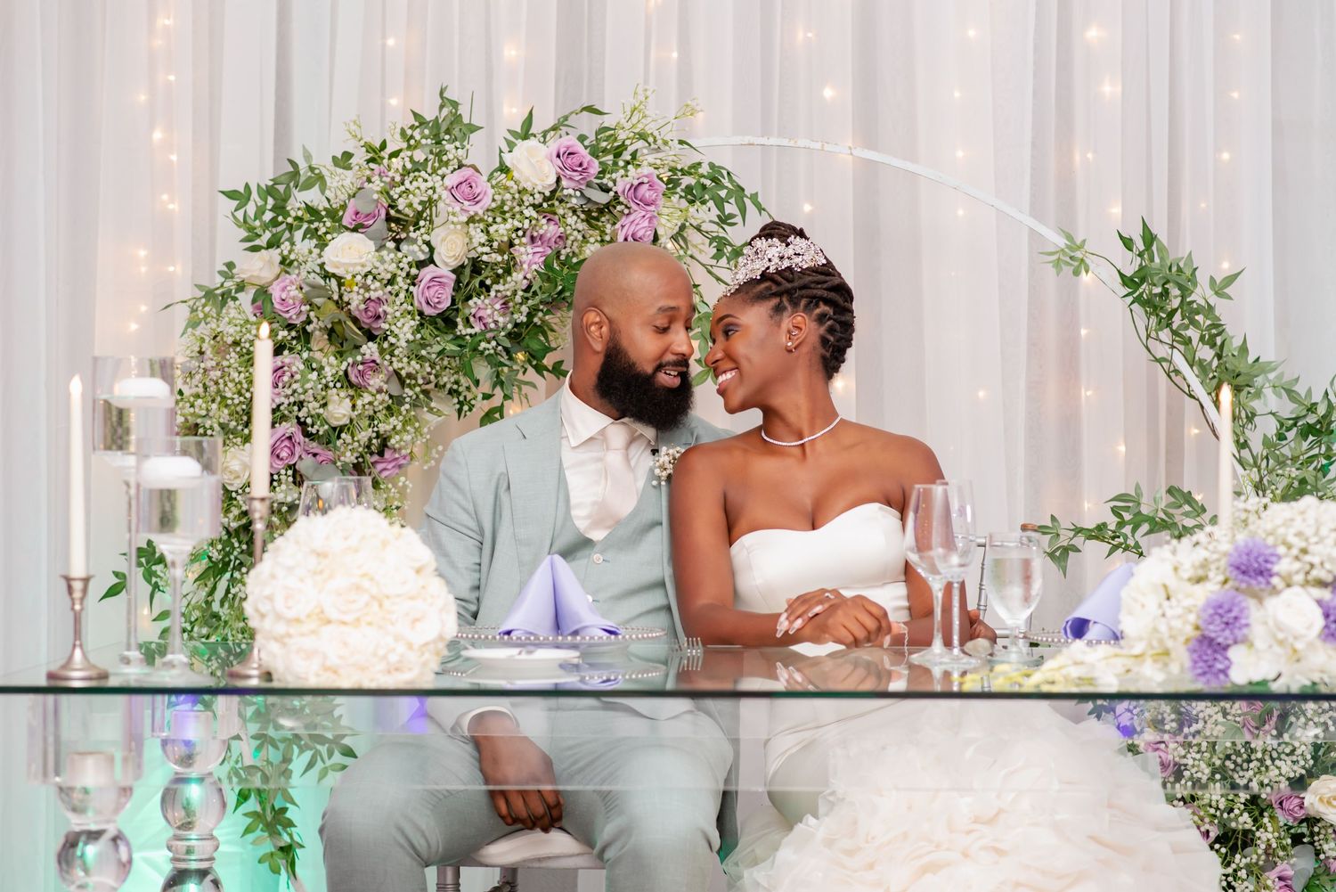 Couple sits at decorated sweetheart table with floral arch and white decor.