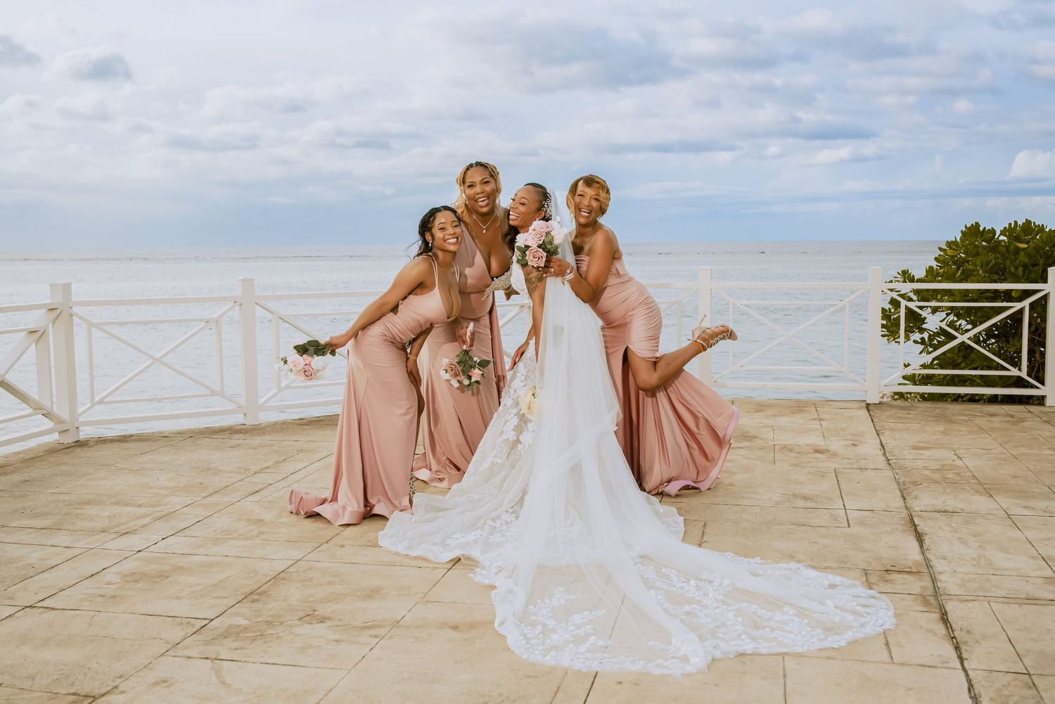 Bridesmaids in pink dresses pose playfully with bride in white gown by the ocean.