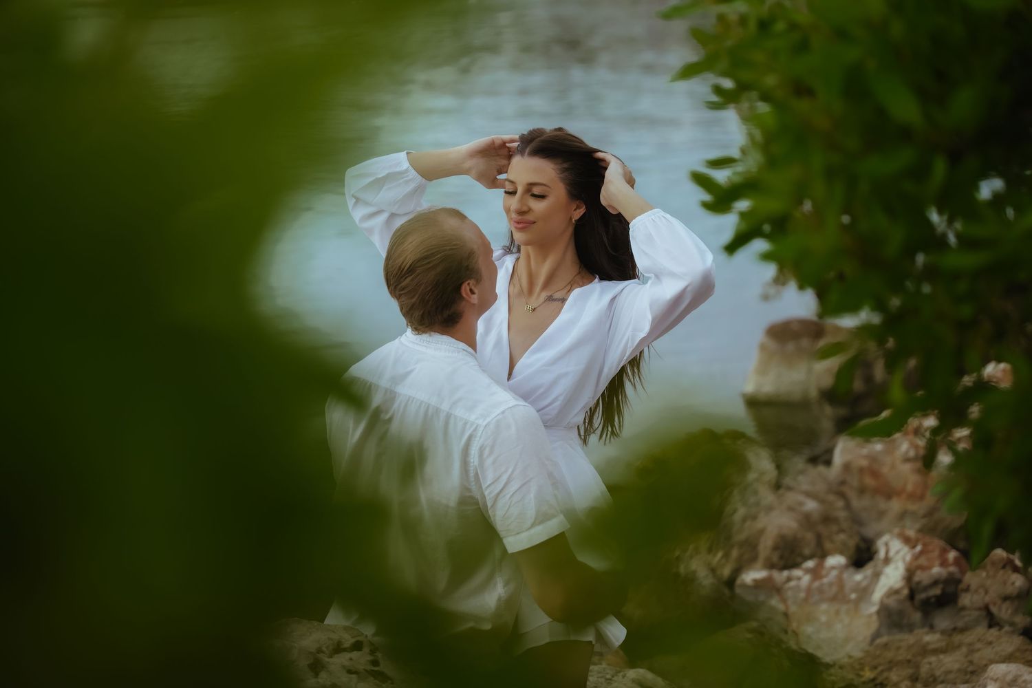 A romantic couple in white outfits share an intimate moment by the water's edge framed through foliage.