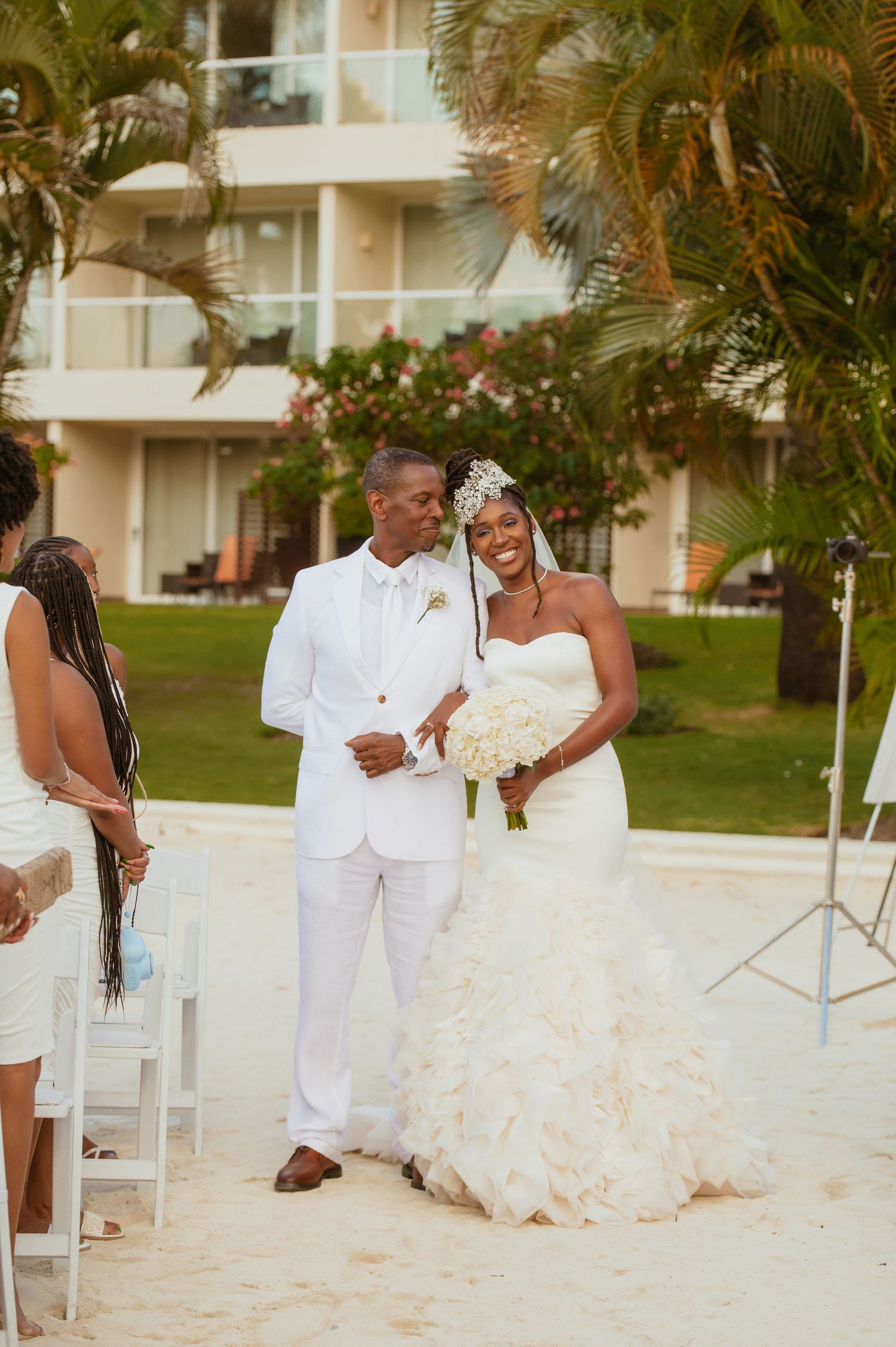 A romantic beach wedding scene with a couple sharing a moment on white chairs surrounded by palm trees.