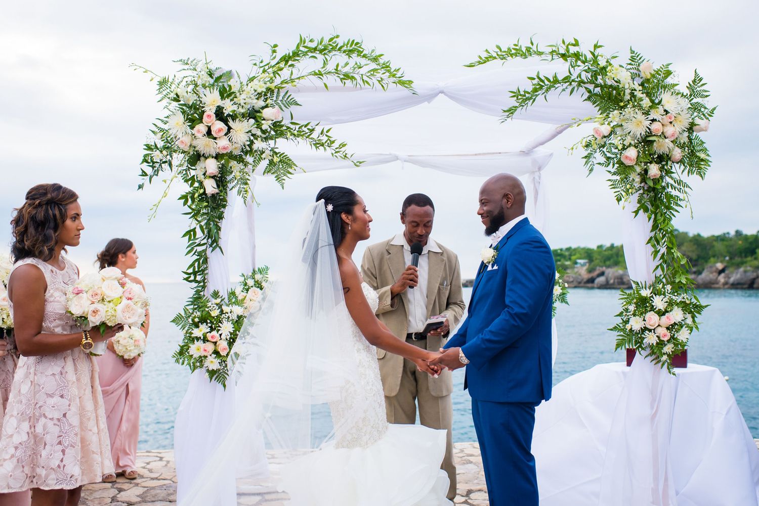 Waterfront wedding ceremony under white floral arch with pink and cream flowers.