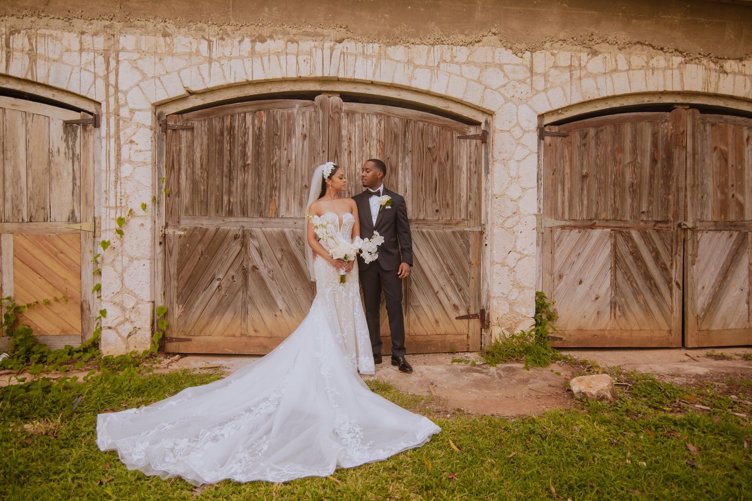 Elegant wedding couple poses against stone wall backdrop in romantic garden setting.