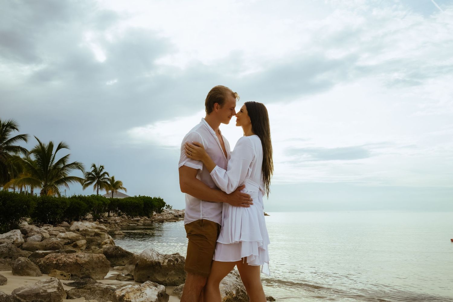 A couple embraces on a rocky tropical shoreline with palm trees and ocean views.