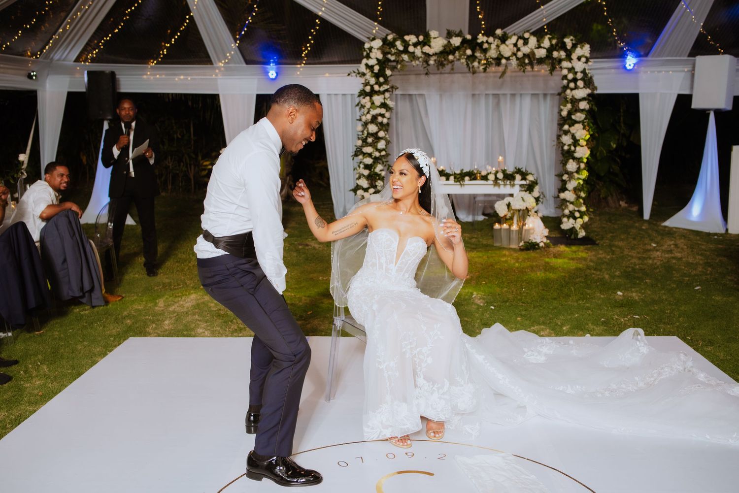 Couple performs energetic dance moves on white dance floor during wedding reception.