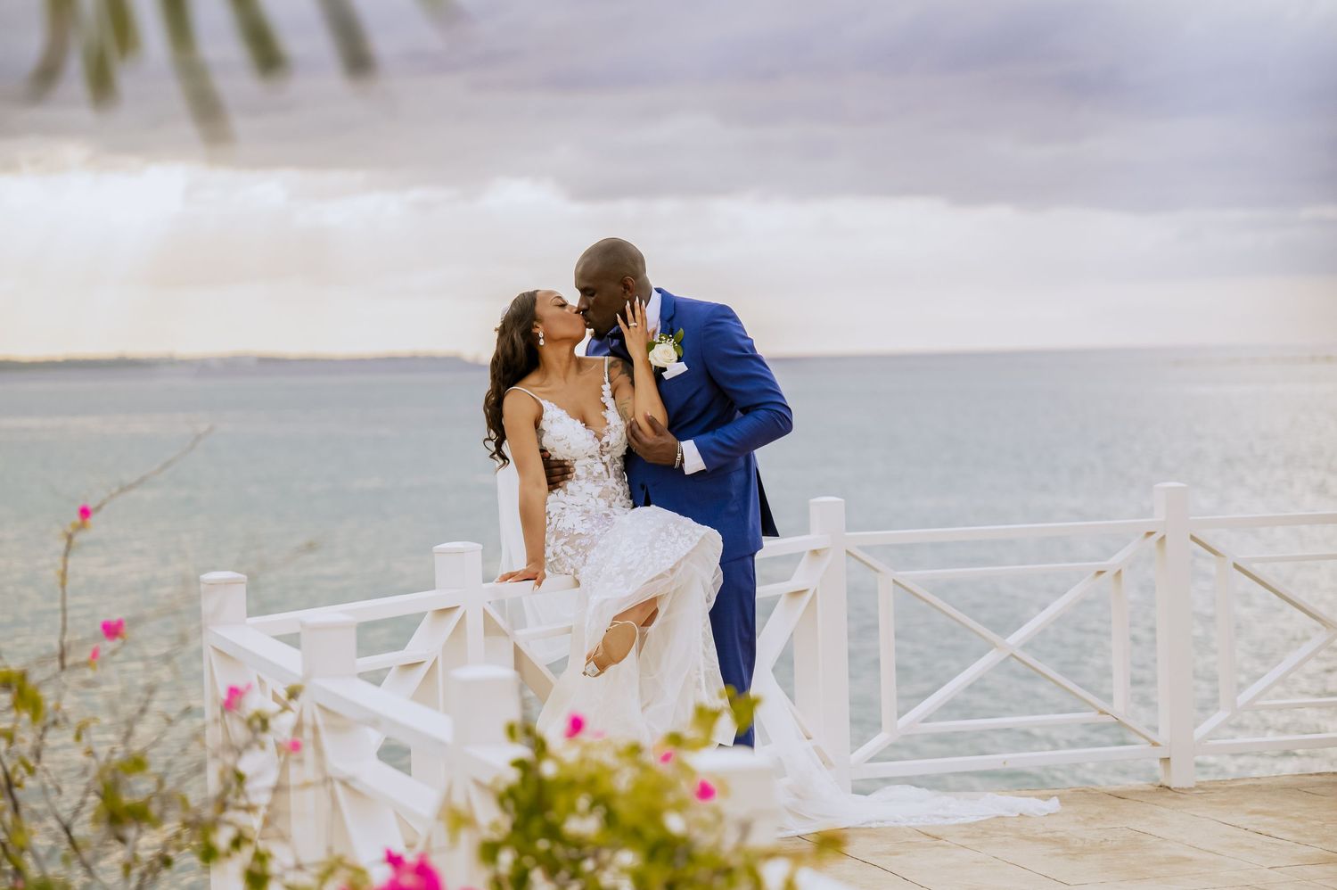 Wedding couple embraces on oceanfront deck with tropical flowers and scenic views.